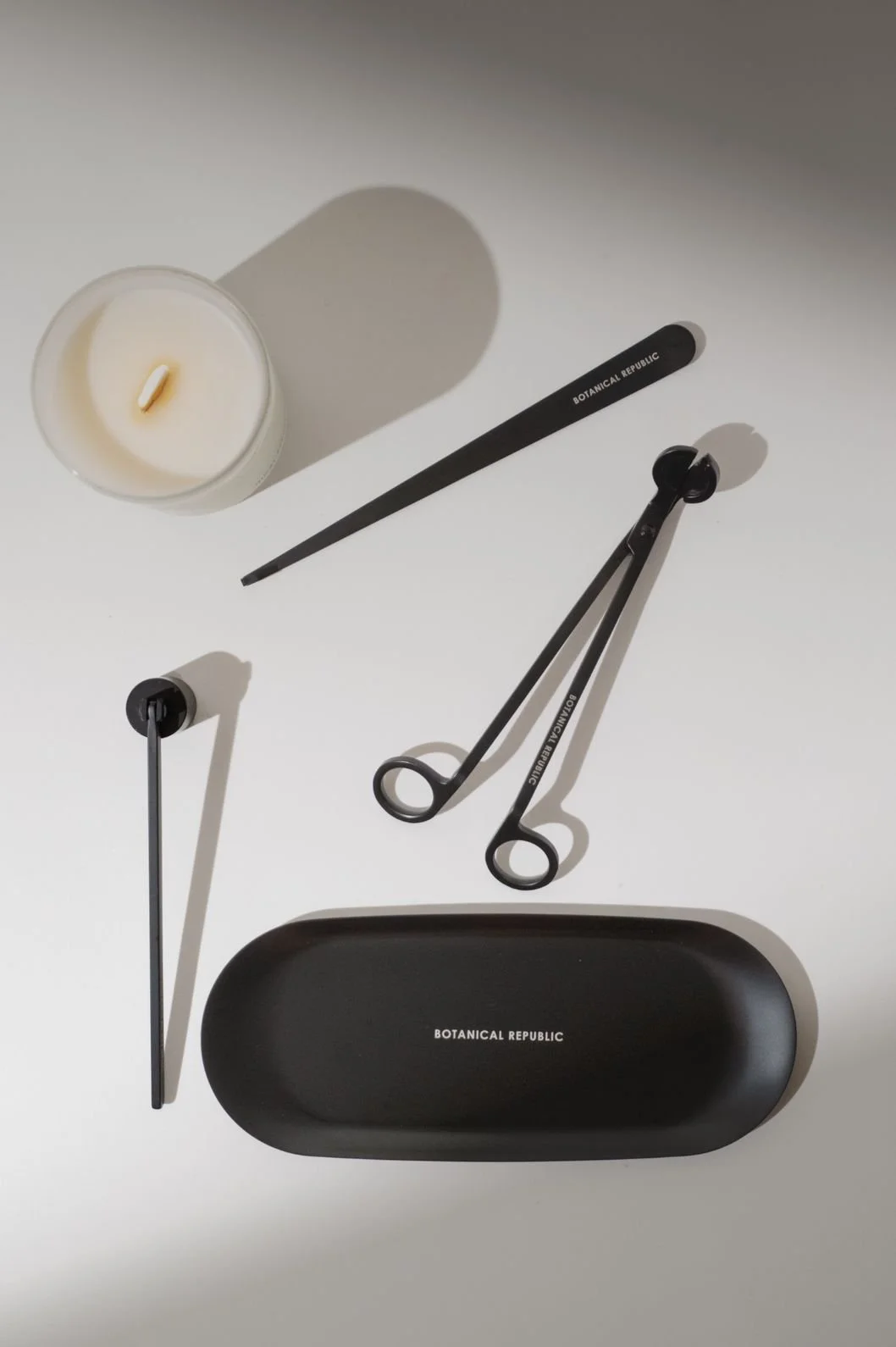 Seattle product photography of a white candle surrounded by black candle accessories such as a candle snuffer, candle trimmer, and wick dipper with dark moody shadows