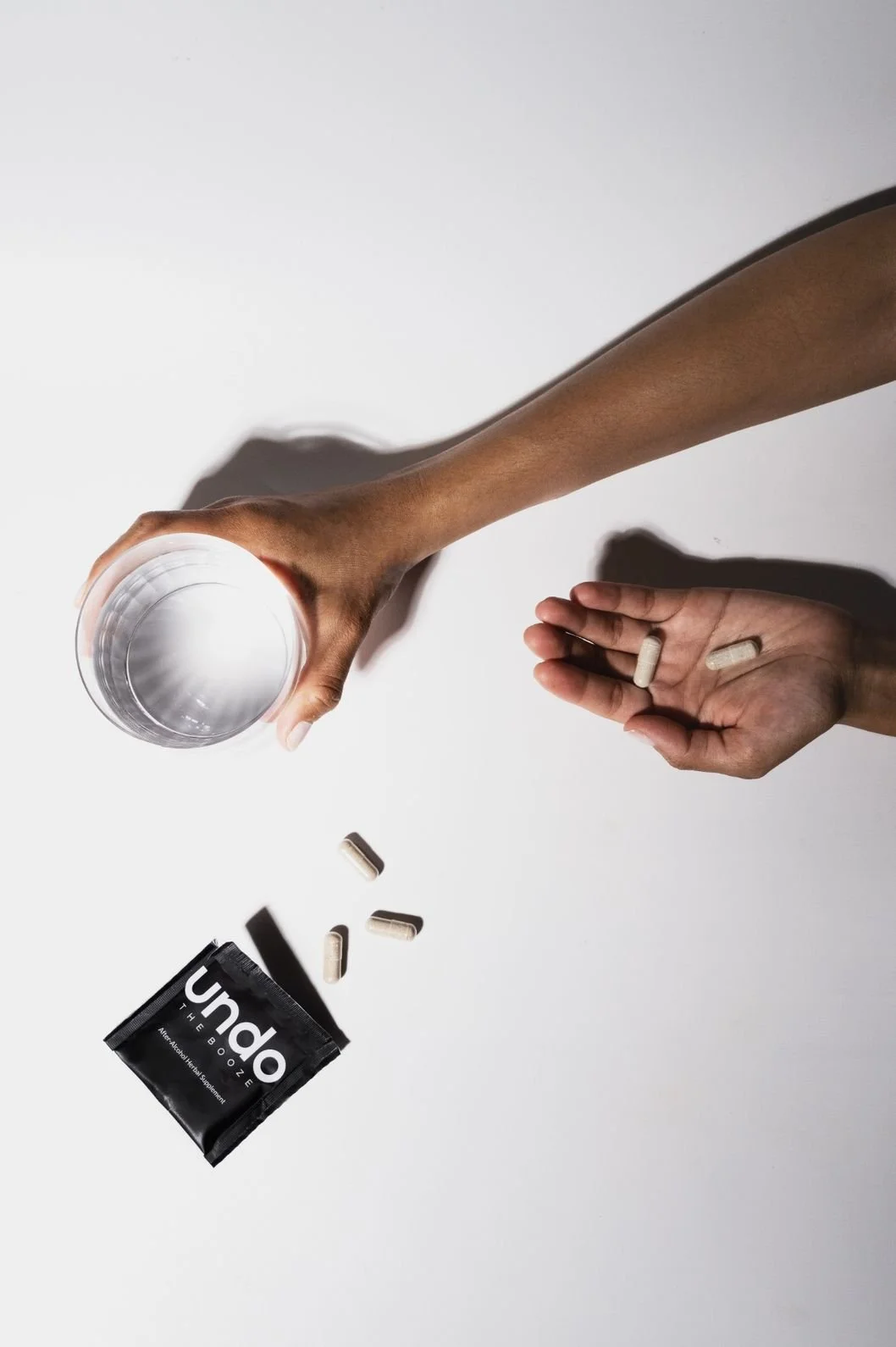 Product photography of a herbal supplement  in an open packet with pills spilling out next to a model's hands - one hand holding a glass of water and the other hand holding two pills