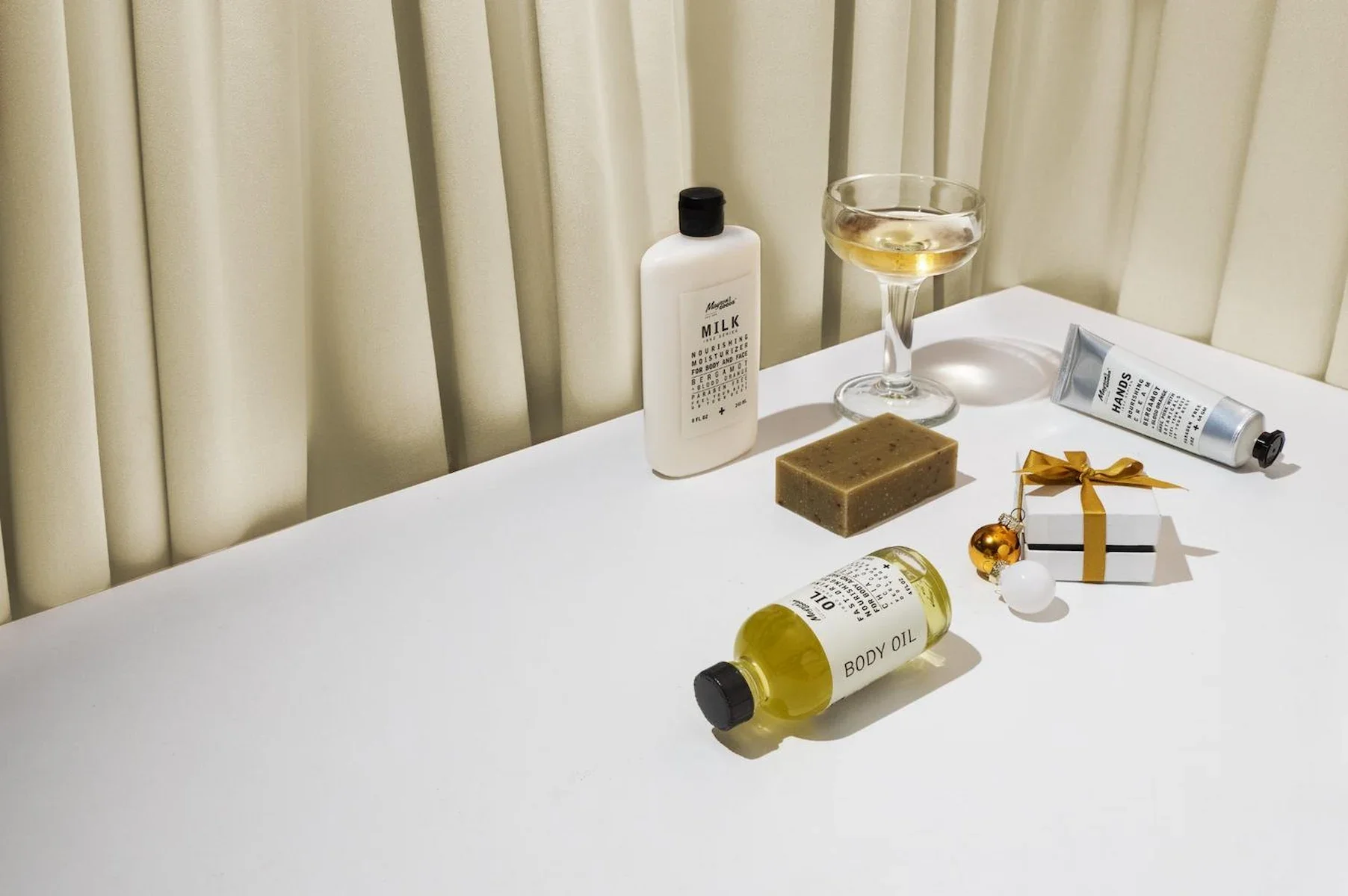 Skincare product photography of a group of products laid out on a white table top with small gifts, holiday ornaments, and a glass of champagne with a gold curtain background