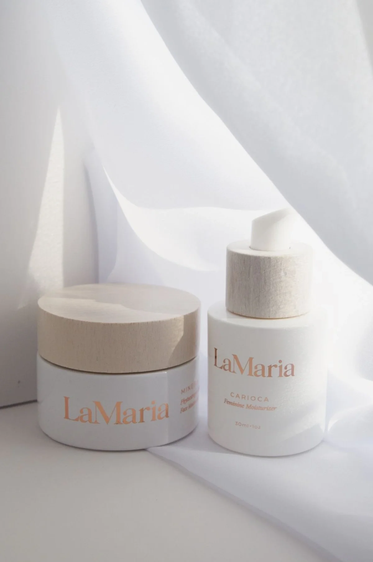 Skincare product photography of a face cream and serum with draped white fabric and soft lighting coming through