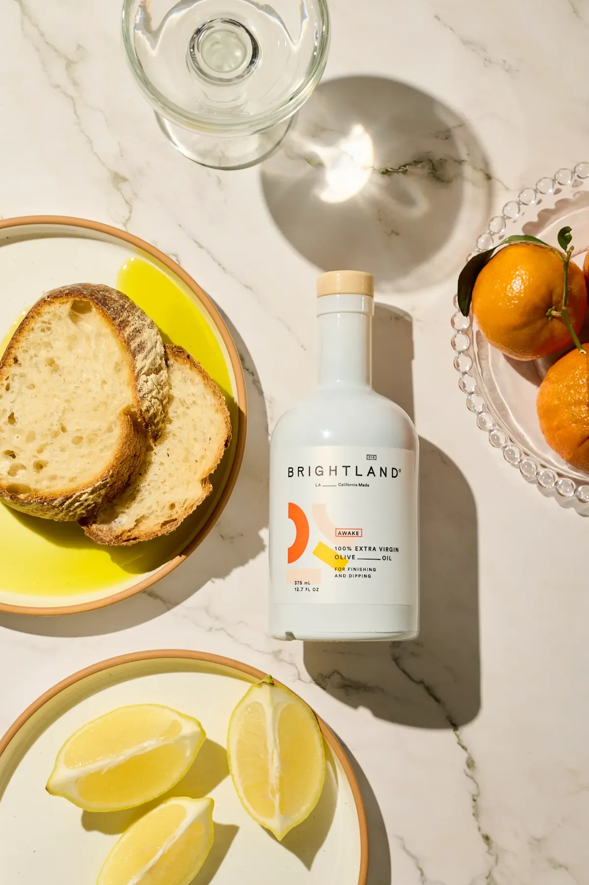 Premium food product photography showcasing an olive oil bottle styled on a marble tabletop, surrounded by fresh bread, lemons, seasonal fruit, and glassware—perfect for gourmet brands, packaging, e-commerce, and editorial food campaigns.