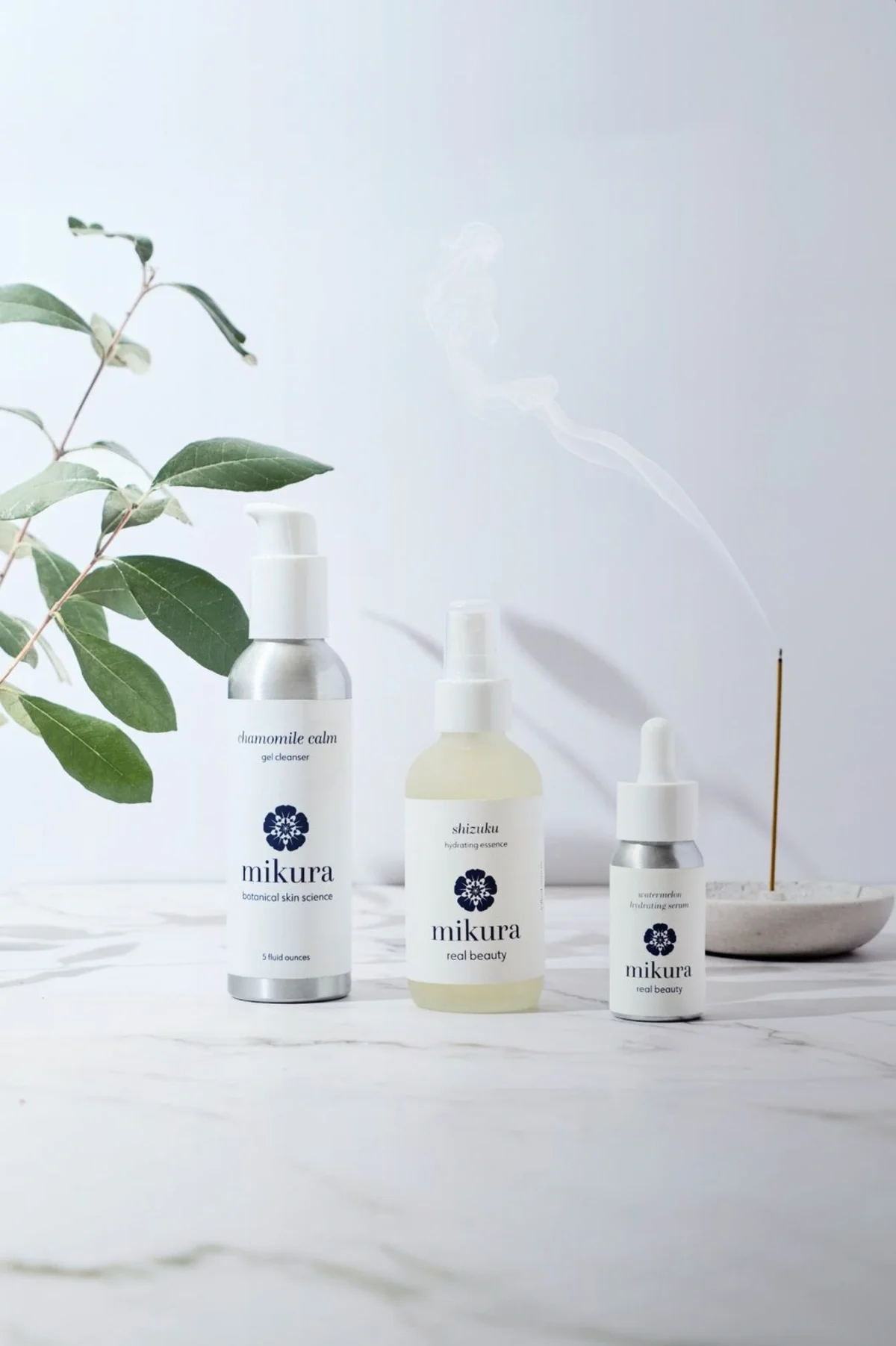 Skincare product photography of a toner, essence, and serum lined up in a row with a green plant and burning incense in the background