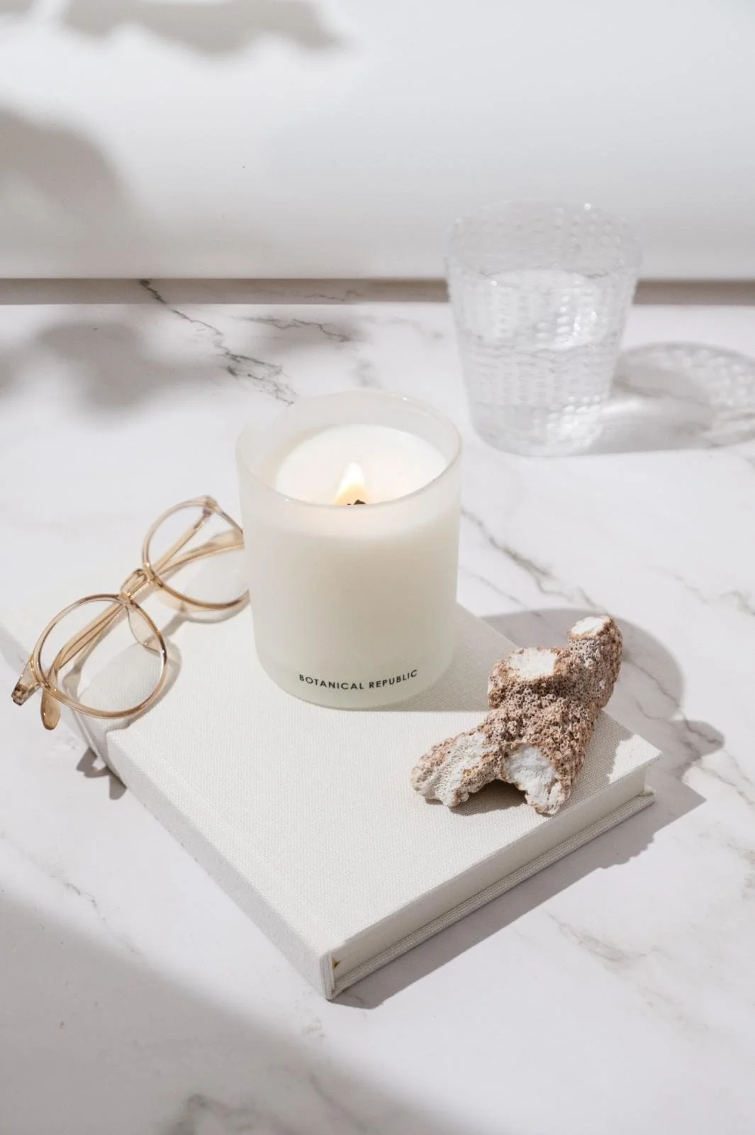 Seattle product photography of a white candle on a book surrounded by a pair of glasses and a piece of coral on a white background