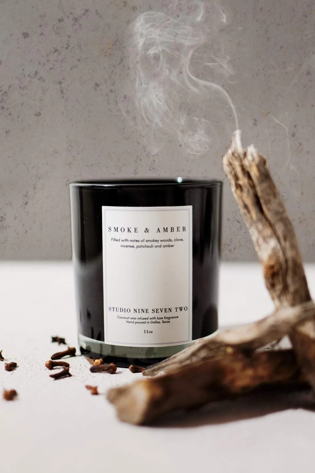 Seattle candle product photography of a black candle surrounded by smoking pieces of wood and cloves on a white tabletop with a grey concrete background