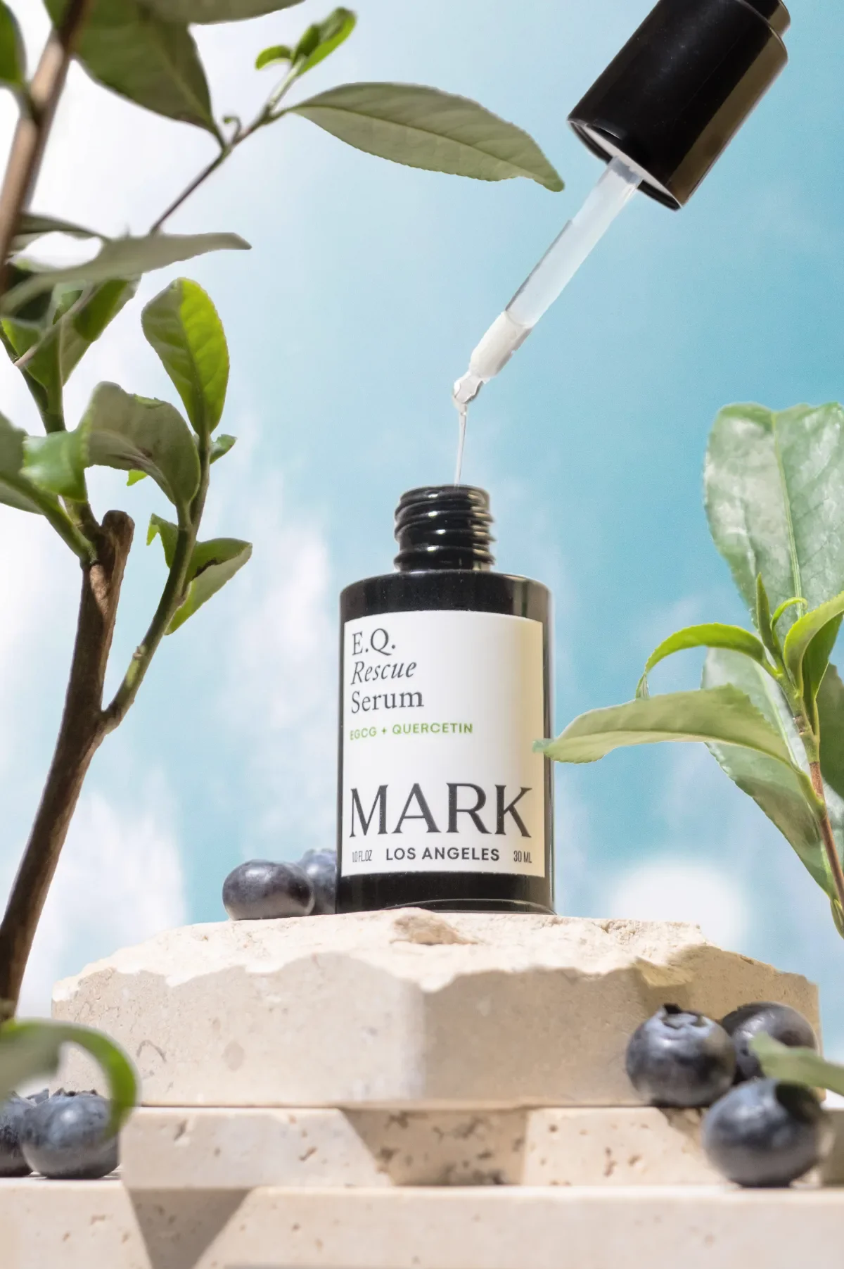 Skincare product photography of a serum bottle set on stone blocks surrounded by blueberries and green plants with a blue sky and the dropper floating out above. Photo for e-commerce, campaigns, and marketing.