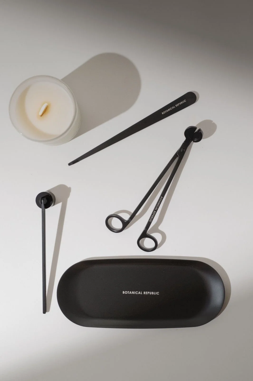 Seattle product photography of a white candle surrounded by black candle accessories such as a candle snuffer, candle trimmer, and wick dipper with dark moody shadows