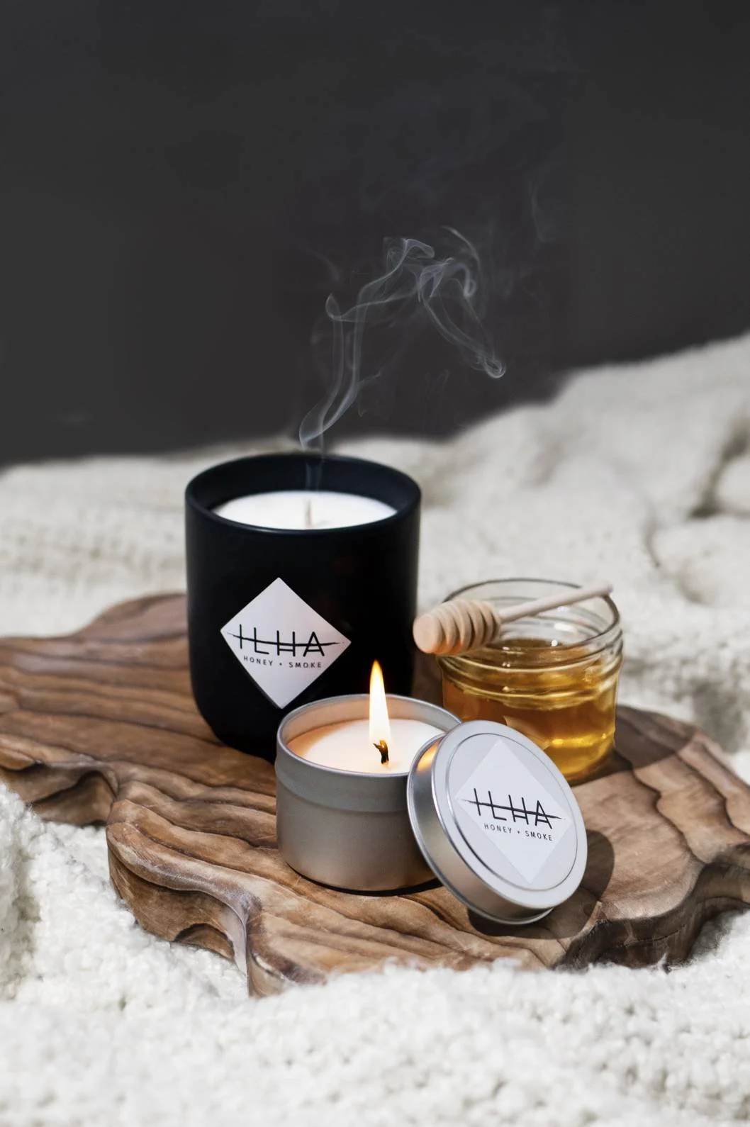 Seattle candle product photography of two candles and a jar of honey on top of a wooden tray on a cozy white blanket against a dark grey background