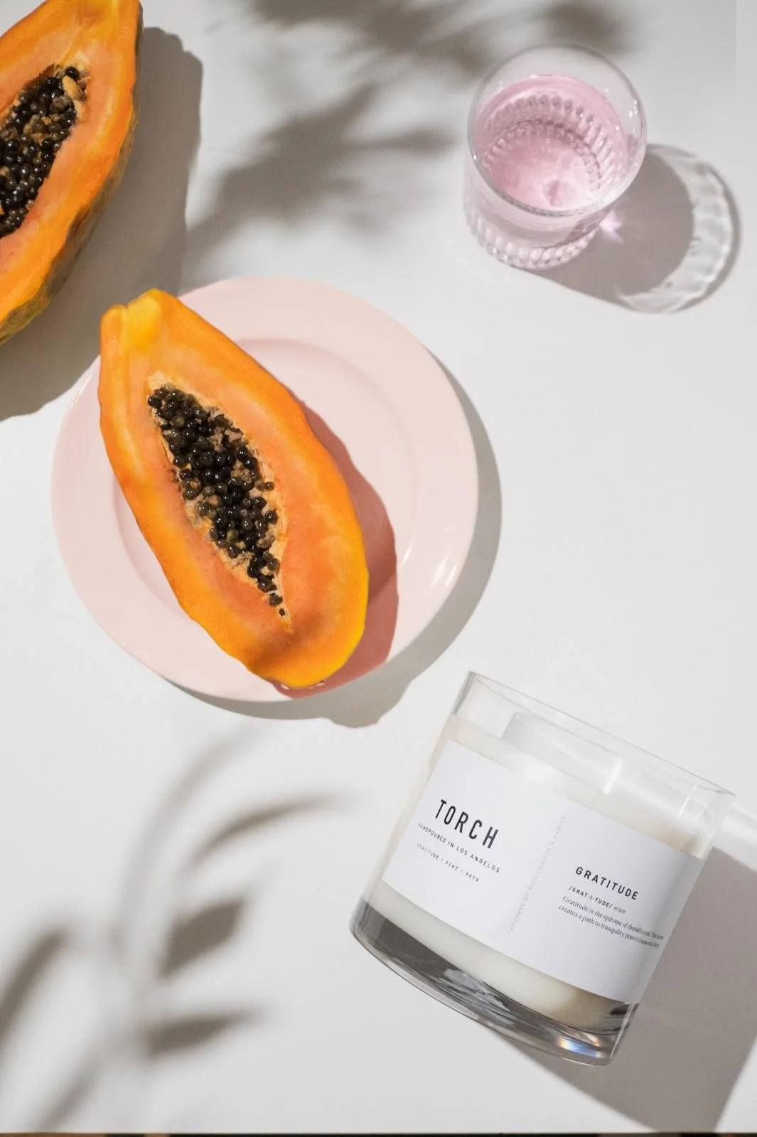 Seattle candle product photography of a white candle on a white tabletop surrounded by cut fresh papaya and a small glass of rose water with soft plant shadows