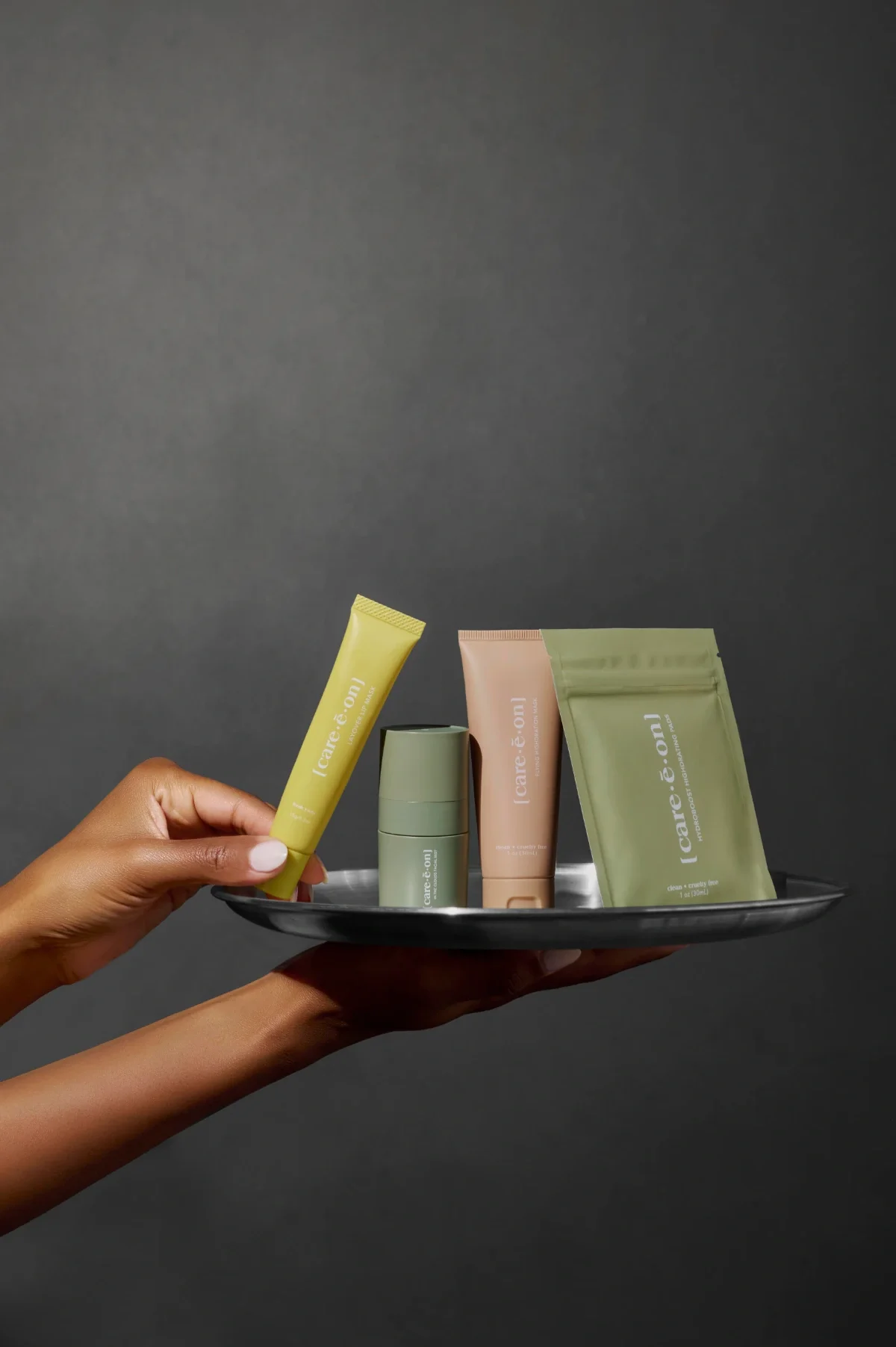 Luxury skincare product photography featuring a hand model holding a sleek silver tray displaying a complete skincare collection, set against a dark gray background with dramatic, moody lighting—ideal for high-end beauty brands, e-commerce, and edito