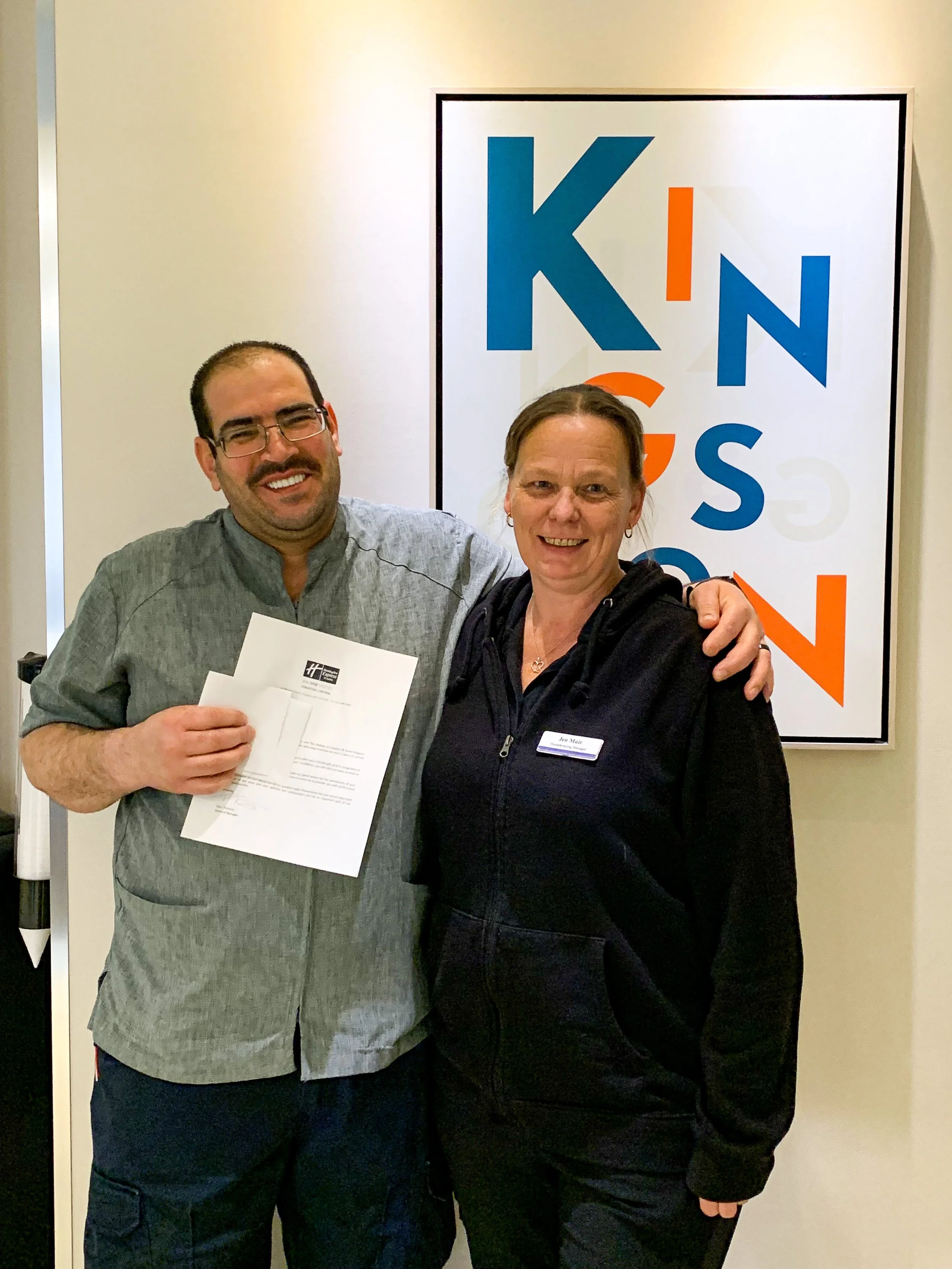 A man and a woman smiling and posing together in front of a large sign that says 'KINGS' with the letters arranged vertically and differently colored. The man is holding some papers, and the woman has her arm around his shoulder.