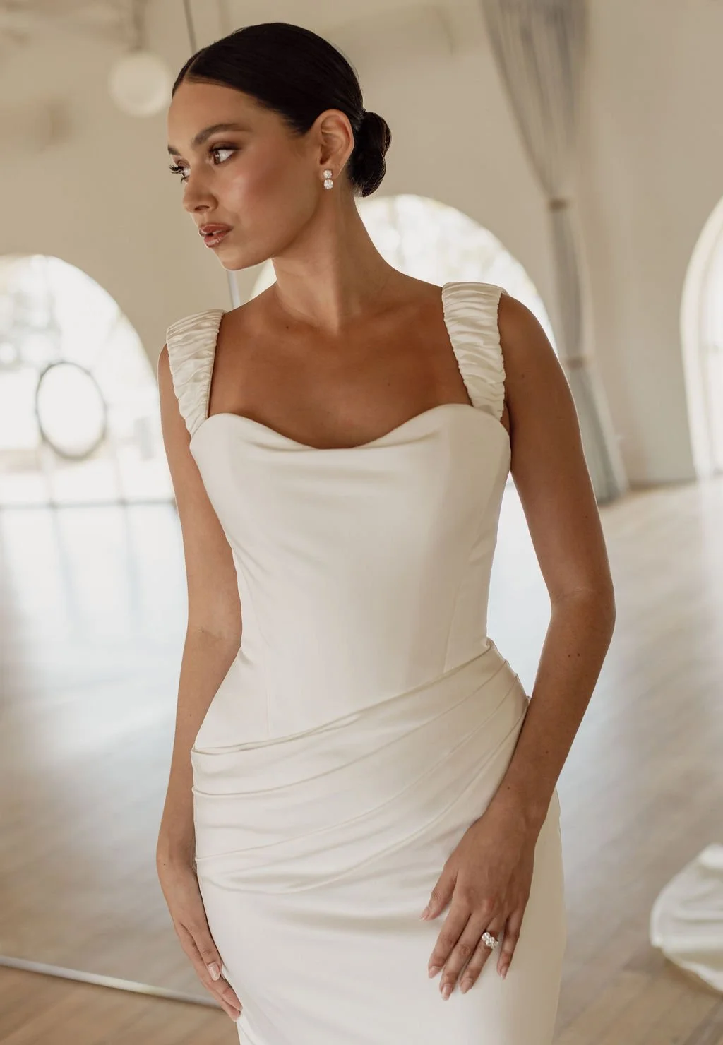 Demi by Trish Peng fitted satin crepe wedding dress featuring an asymmetric draped bodice, off-the-shoulder cowl neckline, removable straps and softly ruched skirt, available at Spellbound Bridal Ireland.
