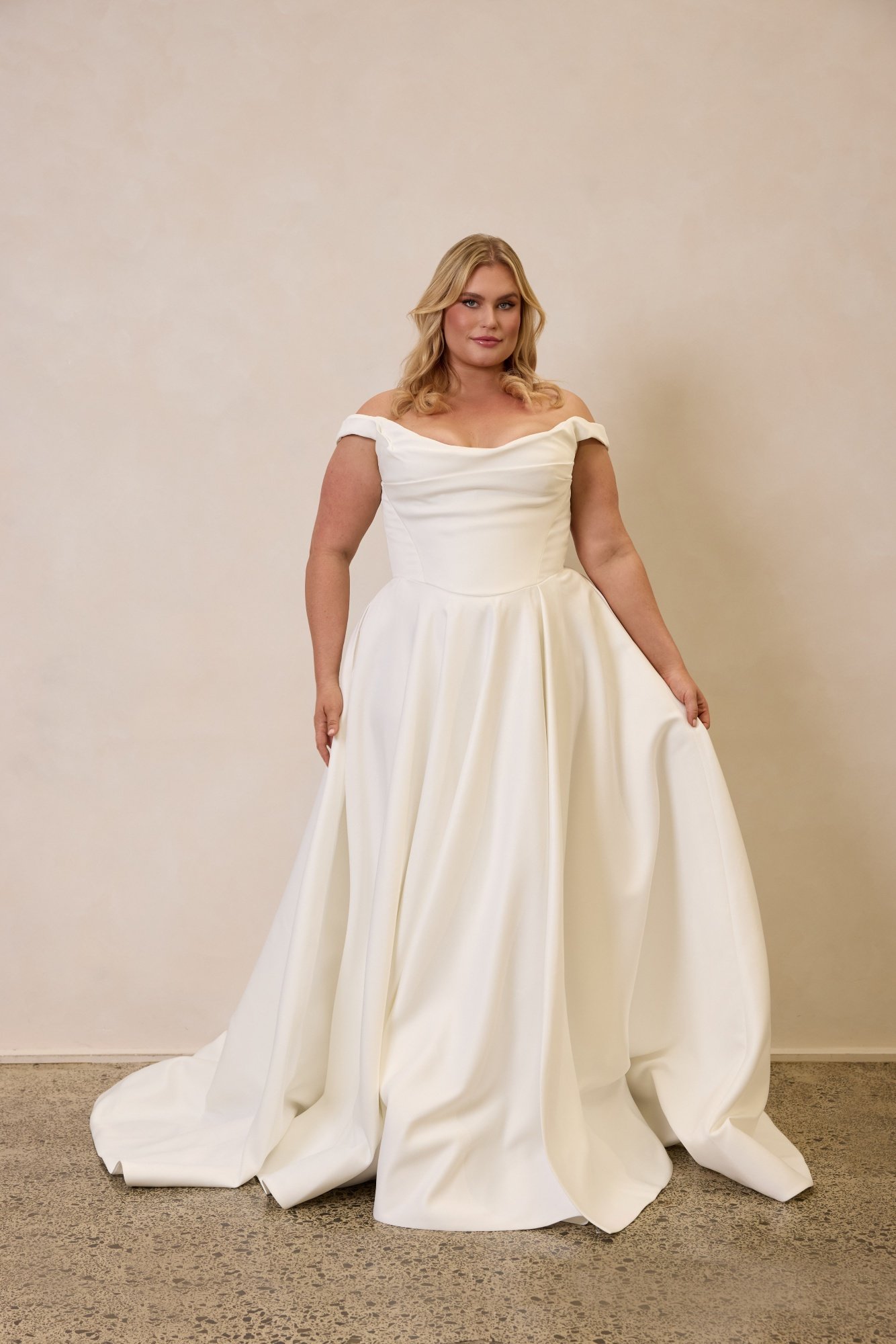 Emily by Trish Peng matte satin wedding dress featuring a draped off-the-shoulder neckline, ruched bodice, sculpted basque waist, A-line skirt and dramatic side split, available in the Curve Collection at Spellbound Bridal Ireland.