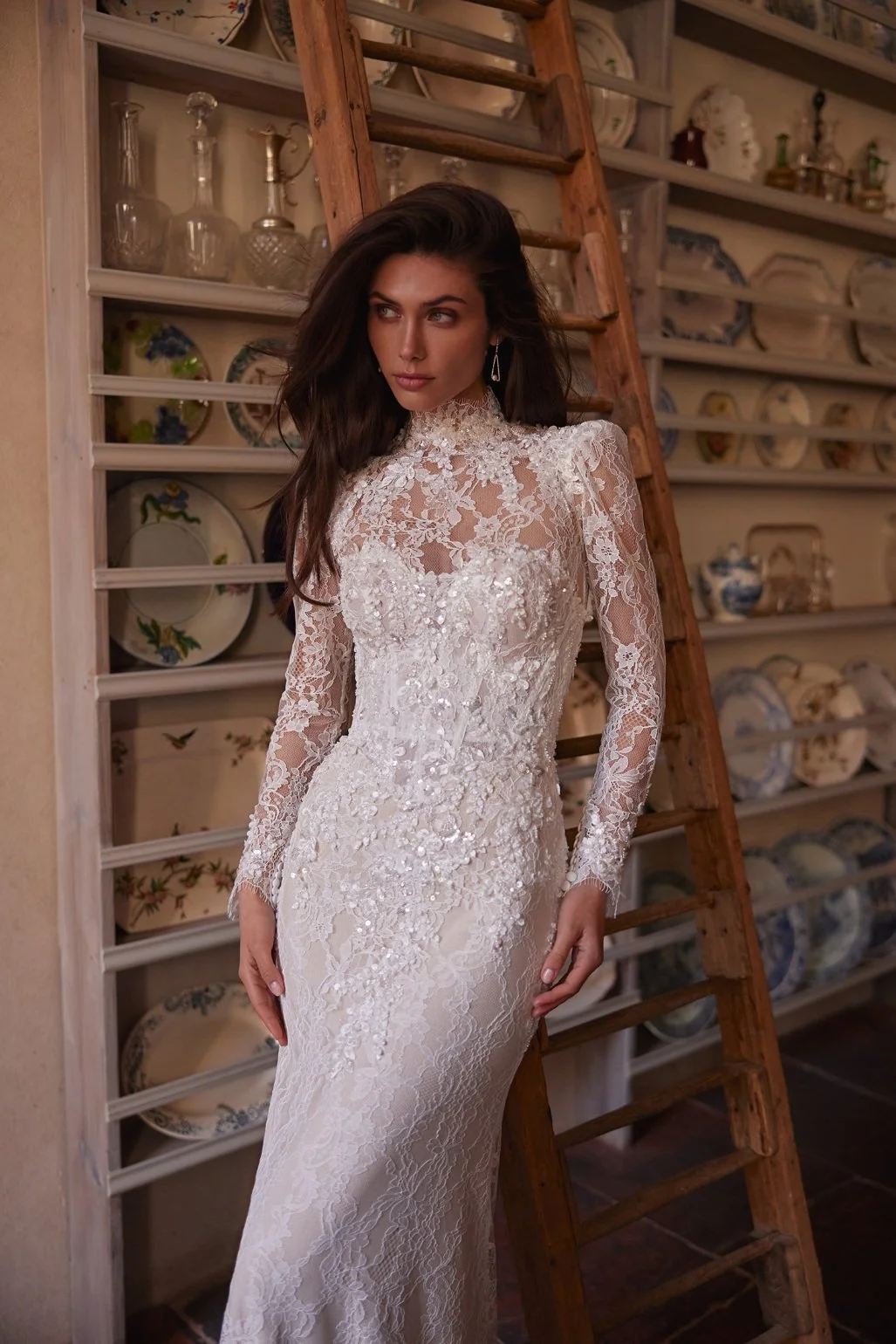 The Lisandra wedding dress is crafted from intricate Alençon lace. This mermaid gown features floral appliqués with delicate bead detailing. A detachable sheer high-neck bolero with long sleeves adds sophistication, while the removable A-line overski