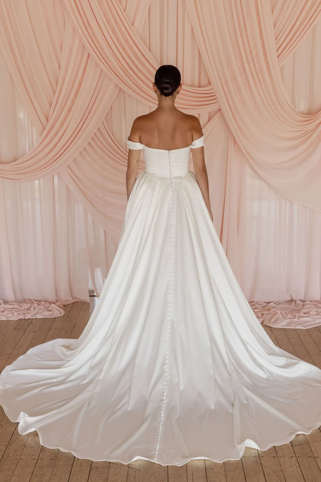 Diana by Trish Peng off-white satin wedding dress with a draped off-the-shoulder neckline, structured basque waist, A-line skirt and detachable bustle, available at Spellbound Bridal Ireland.