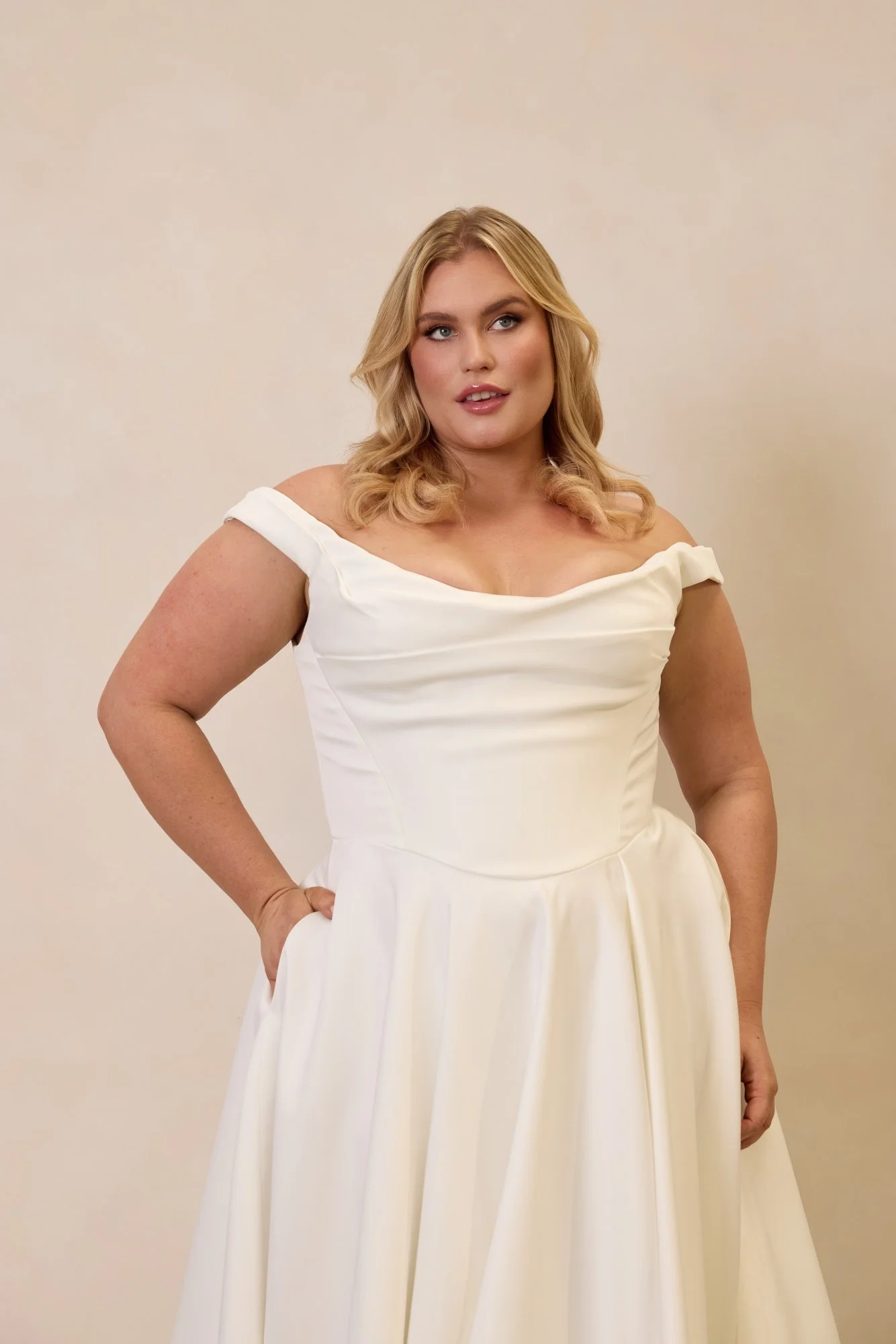 Emily by Trish Peng matte satin wedding dress featuring a draped off-the-shoulder neckline, ruched bodice, sculpted basque waist, A-line skirt and dramatic side split, available in the Curve Collection at Spellbound Bridal Ireland.
