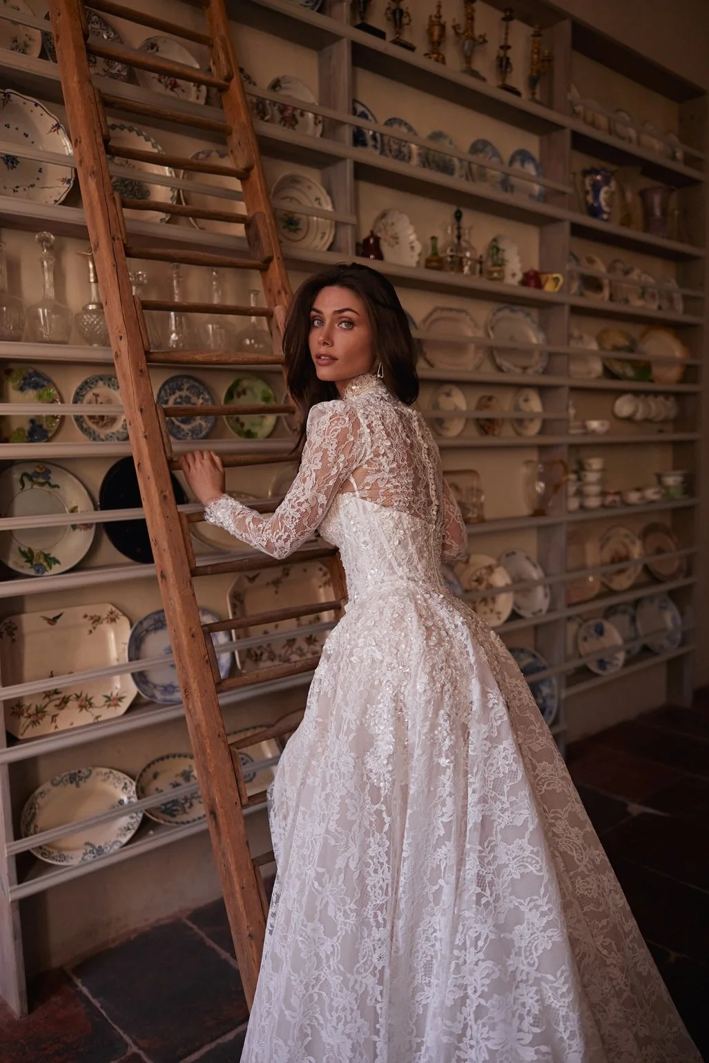 The Lisandra wedding dress is crafted from intricate Alençon lace. This mermaid gown features floral appliqués with delicate bead detailing. A detachable sheer high-neck bolero with long sleeves adds sophistication, while the removable A-line overski