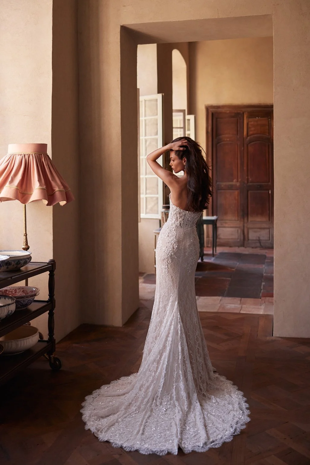 The Lisandra wedding dress is crafted from intricate Alençon lace. This mermaid gown features floral appliqués with delicate bead detailing. A detachable sheer high-neck bolero with long sleeves adds sophistication, while the removable A-line overski