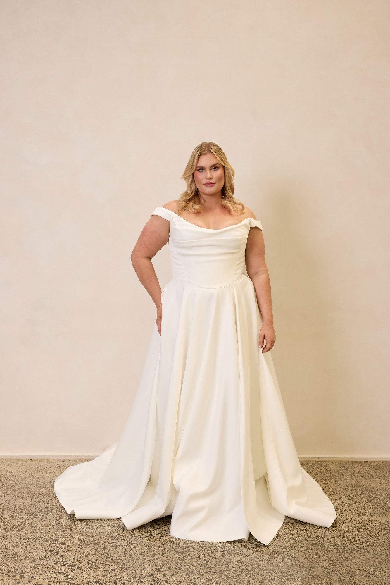 Emily by Trish Peng matte satin wedding dress featuring a draped off-the-shoulder neckline, ruched bodice, sculpted basque waist, A-line skirt and dramatic side split, available in the Curve Collection at Spellbound Bridal Ireland.