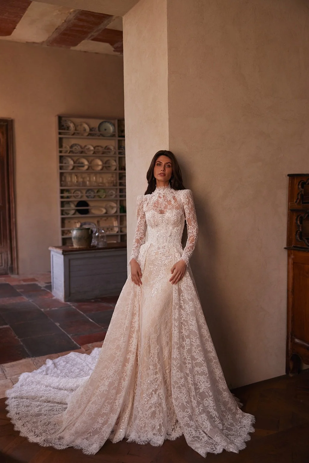 The Lisandra wedding dress is crafted from intricate Alençon lace. This mermaid gown features floral appliqués with delicate bead detailing. A detachable sheer high-neck bolero with long sleeves adds sophistication, while the removable A-line overski
