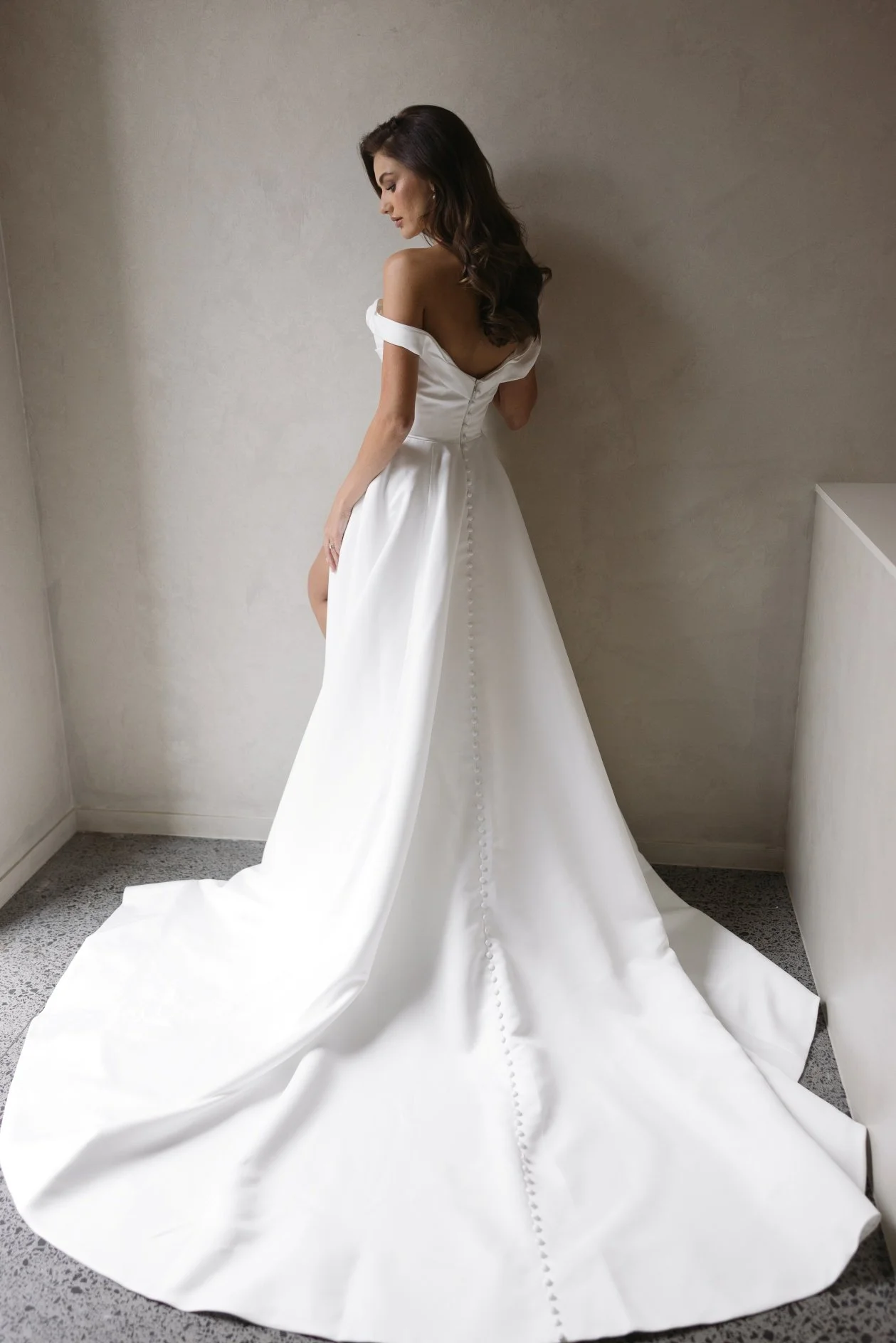 Emily by Trish Peng matte satin wedding dress featuring a draped off-the-shoulder neckline, ruched bodice, sculpted basque waist, A-line skirt and dramatic side split, available at Spellbound Bridal Ireland.

