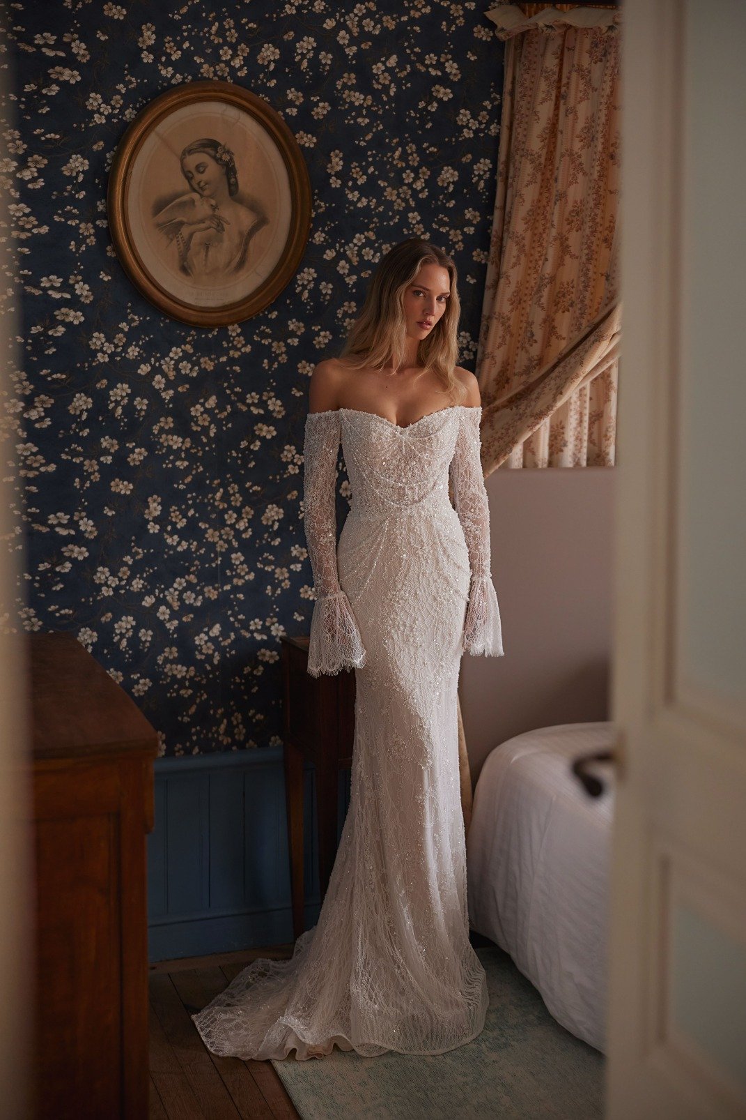 KATHERINE is a mermaid gown made of Chantilly lace and embellished with crystals and pearls. The bodice is decorated with draped strings of pearls. The off the shoulder long lace sleeves finish with a flutter cuff. A delicate detachable skirt adds a