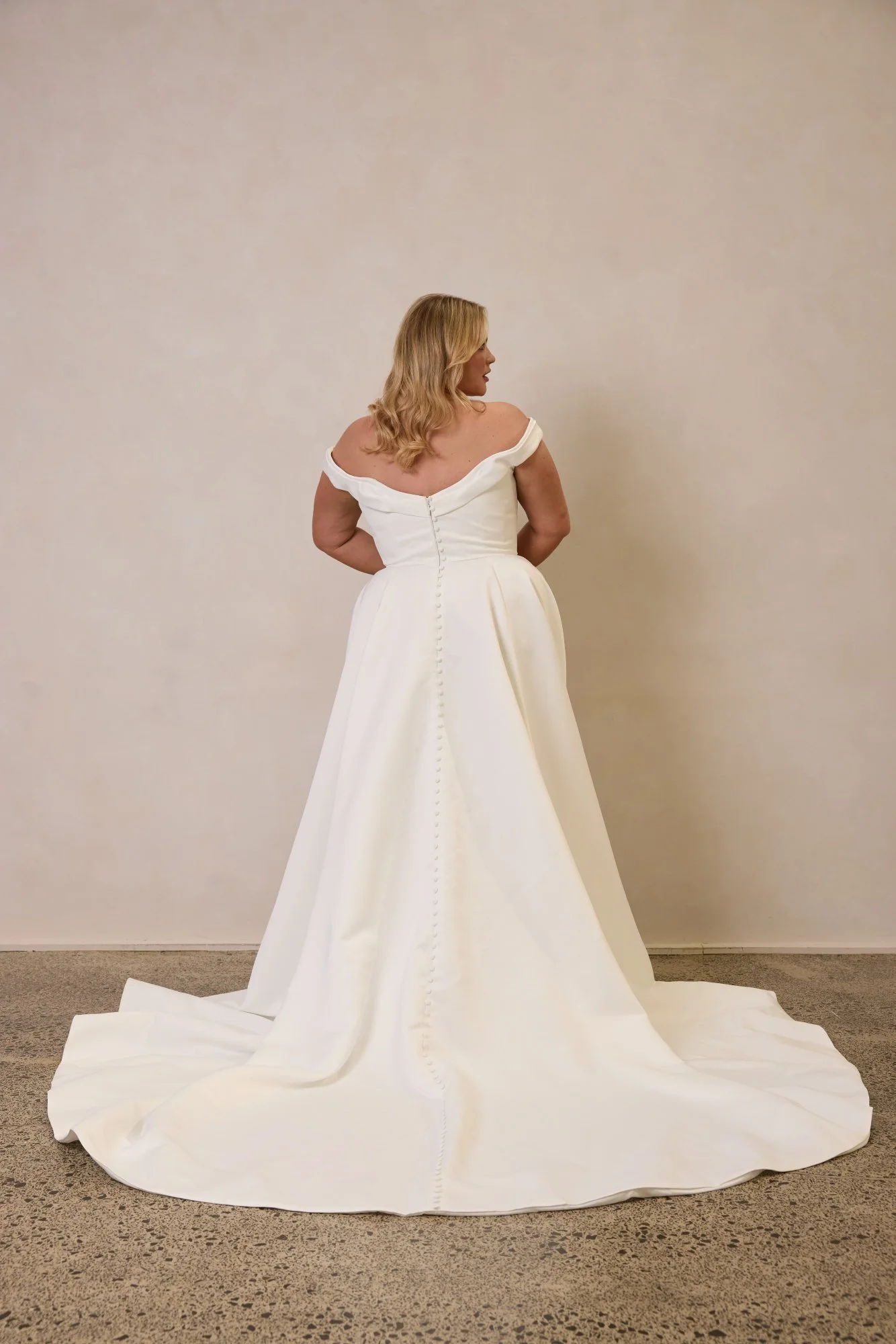 Emily by Trish Peng matte satin wedding dress featuring a draped off-the-shoulder neckline, ruched bodice, sculpted basque waist, A-line skirt and dramatic side split, available in the Curve Collection at Spellbound Bridal Ireland.