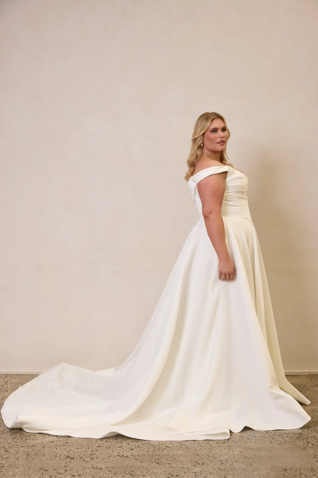 Emily by Trish Peng matte satin wedding dress featuring a draped off-the-shoulder neckline, ruched bodice, sculpted basque waist, A-line skirt and dramatic side split, available in the Curve Collection at Spellbound Bridal Ireland.