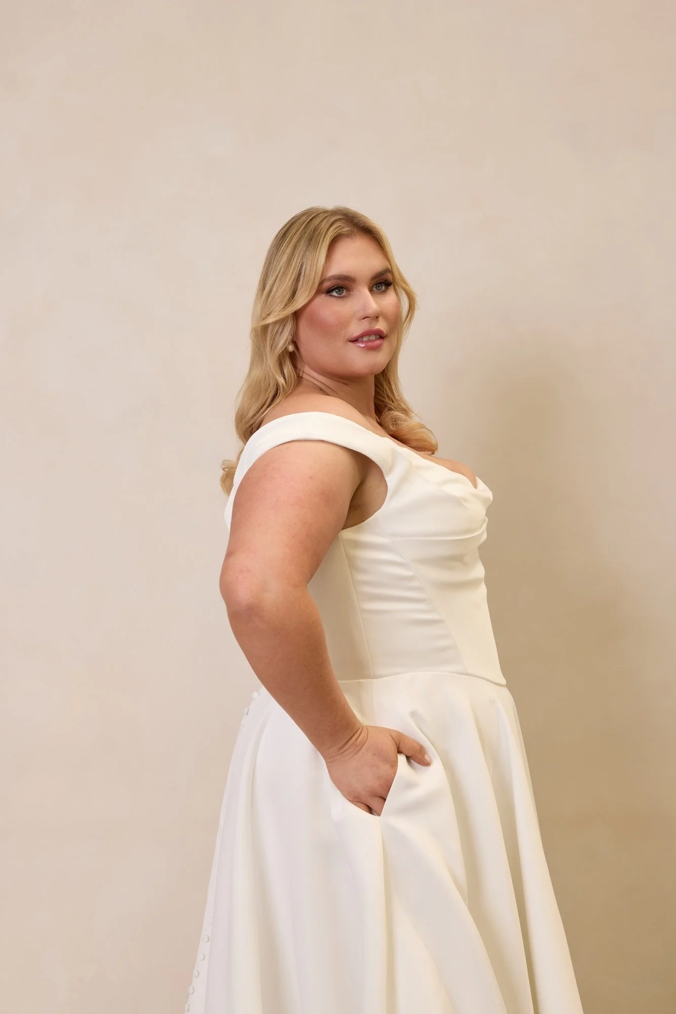 Emily by Trish Peng matte satin wedding dress featuring a draped off-the-shoulder neckline, ruched bodice, sculpted basque waist, A-line skirt and dramatic side split, available in the Curve Collection at Spellbound Bridal Ireland.
