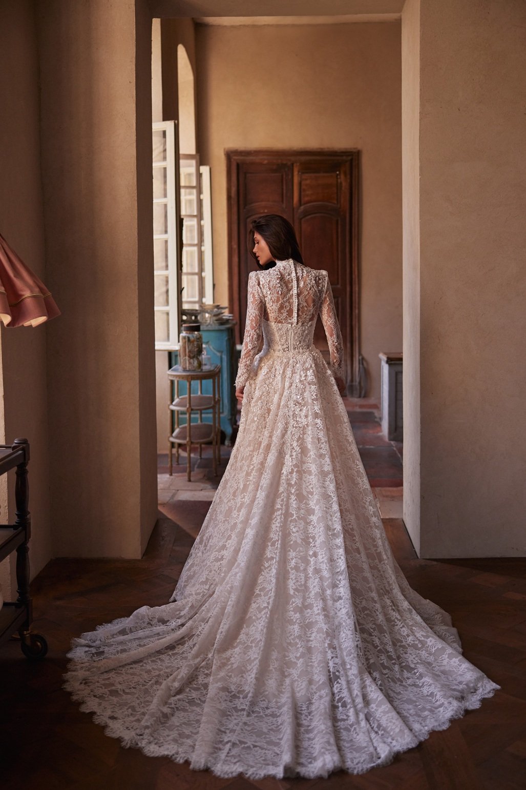 The Lisandra wedding dress is crafted from intricate Alençon lace. This mermaid gown features floral appliqués with delicate bead detailing. A detachable sheer high-neck bolero with long sleeves adds sophistication, while the removable A-line overski