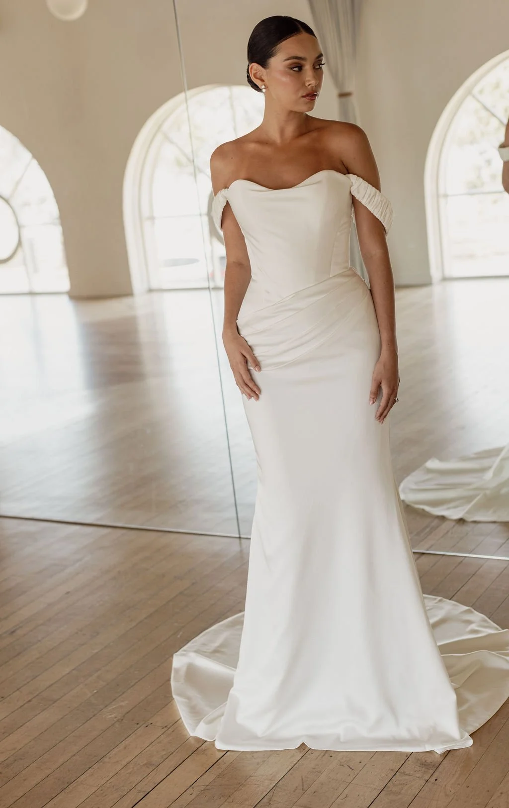 Demi by Trish Peng fitted satin crepe wedding dress featuring an asymmetric draped bodice, off-the-shoulder cowl neckline, removable straps and softly ruched skirt, available at Spellbound Bridal Ireland.

