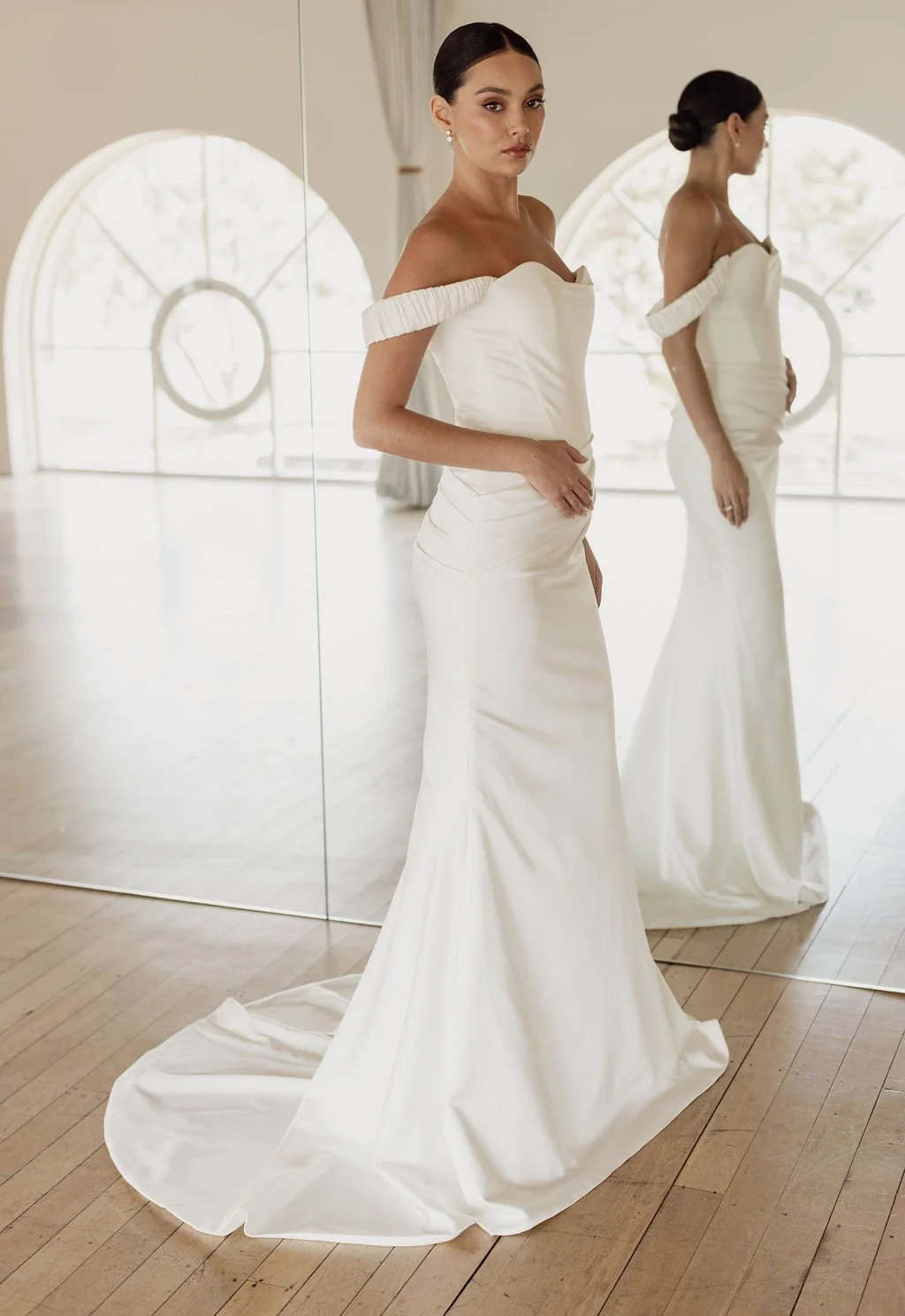 Demi by Trish Peng fitted satin crepe wedding dress featuring an asymmetric draped bodice, off-the-shoulder cowl neckline, removable straps and softly ruched skirt, available at Spellbound Bridal Ireland.
