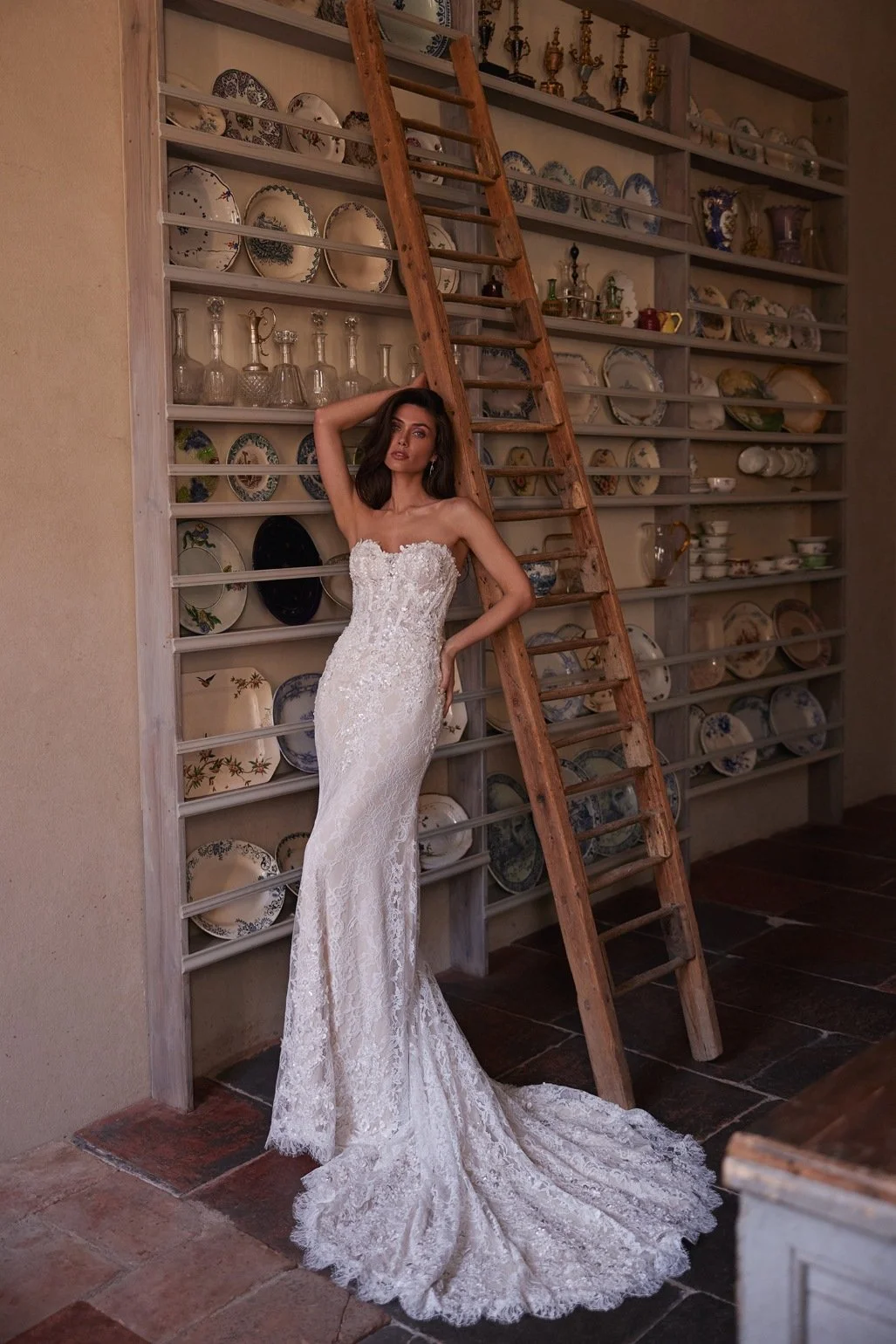 The Lisandra wedding dress is crafted from intricate Alençon lace. This mermaid gown features floral appliqués with delicate bead detailing. A detachable sheer high-neck bolero with long sleeves adds sophistication, while the removable A-line overski