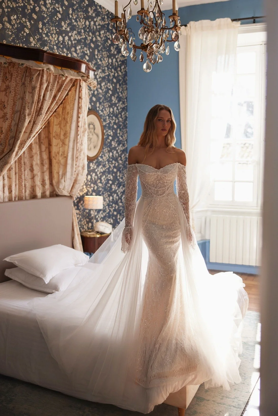 KATHERINE is a mermaid gown made of Chantilly lace and embellished with crystals and pearls. The bodice is decorated with draped strings of pearls. The off the shoulder long lace sleeves finish with a flutter cuff. A delicate detachable skirt adds a