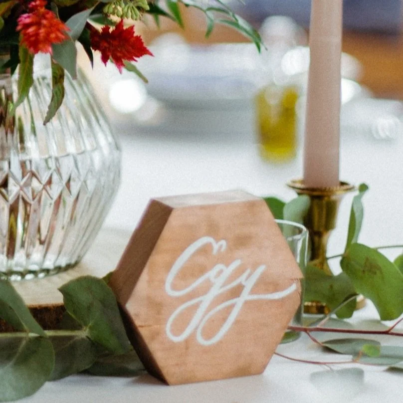 Fából készült hatszögletű tábla, zöld levelekkel körülvéve, egy asztalon, a háttérben egy virágokkal teli üvegvázával és egy sárgaréz tartóban lévő rózsaszín gyertyával.