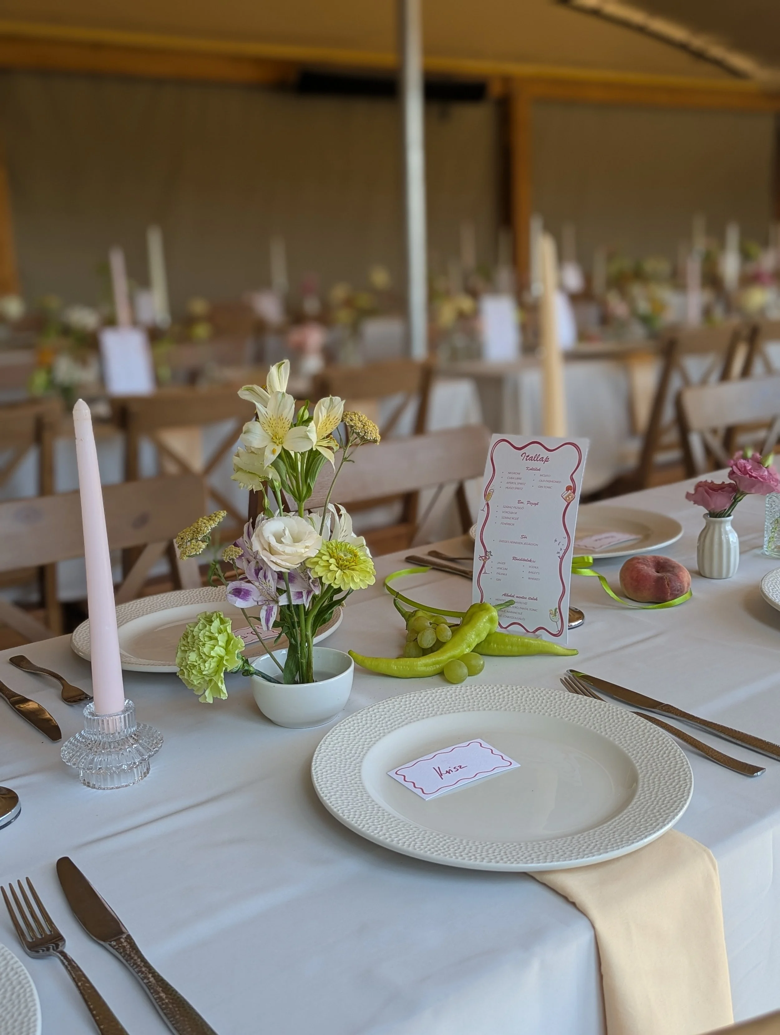 Modern esküvői asztaldekoráció fehér terítővel, pasztell virágkompozíciókkal, kézzel írt ültetőkártyával és gyümölcsös dekor elemekkel. letisztult, elegáns esküvői dekoráció MONA Design stílus
