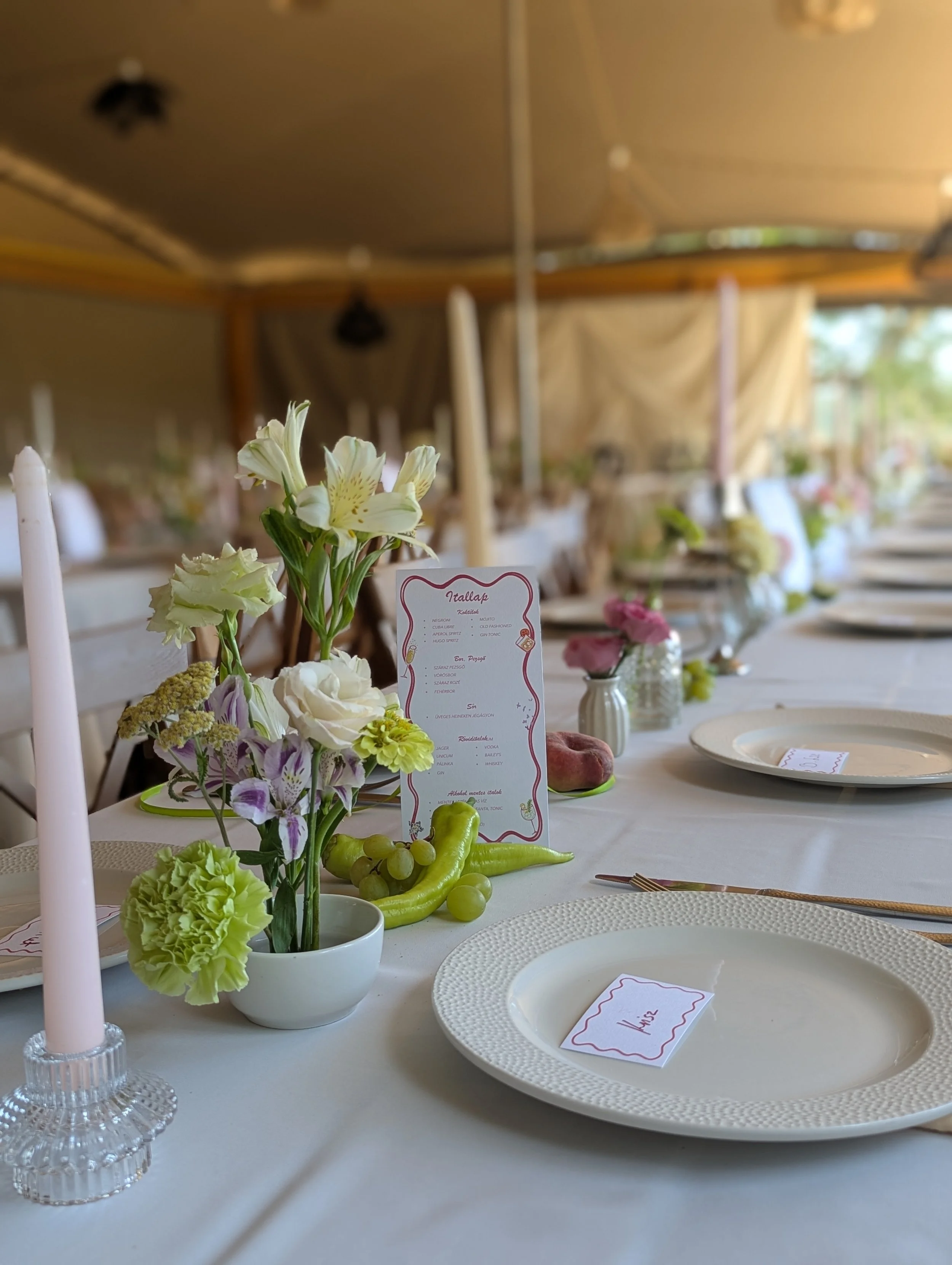 Modern esküvői asztaldekoráció fehér terítővel, pasztell virágkompozíciókkal, kézzel írt ültetőkártyával és gyümölcsös dekor elemekkel. letisztult, elegáns esküvői dekoráció MONA Design stílus