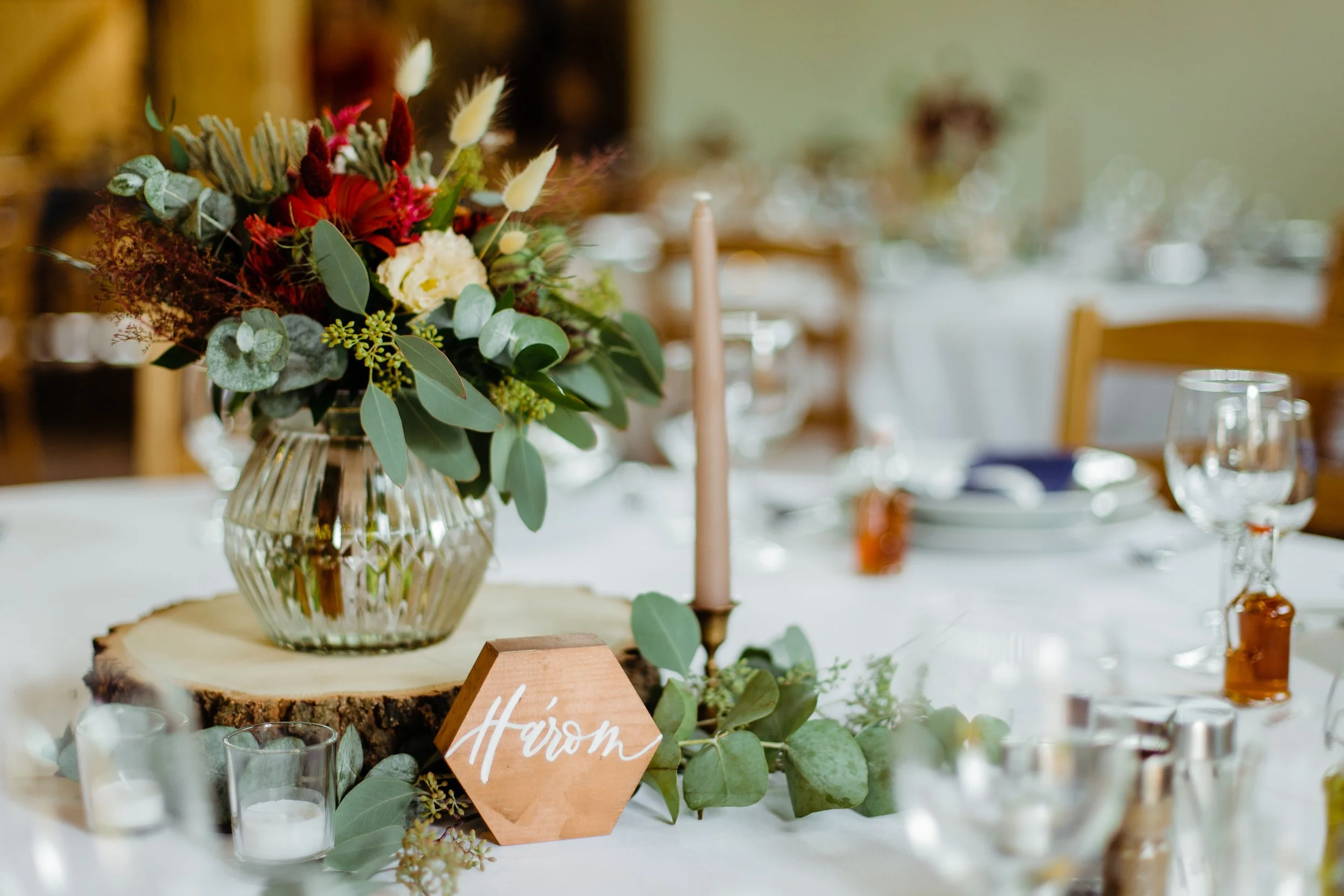 Esküvői vendégasztal dísze vegyes növényekkel és virágokkal díszített üvegvázával, magas rózsaszín gyertyával gyertyatartóban, piros étlappal és egy hatszögletű, fából készült asztalszámmal.
