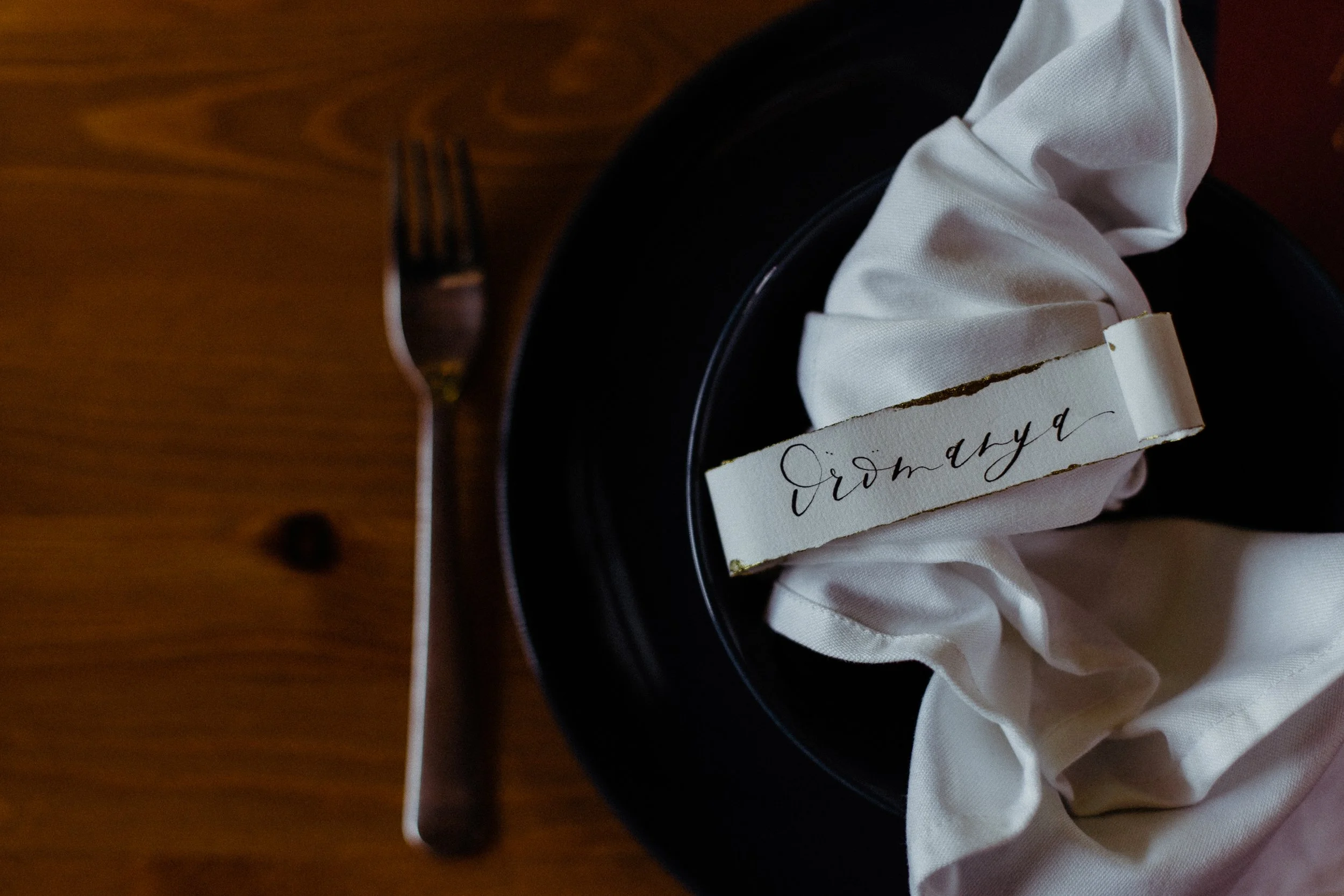 Egy fekete tányér fehér szövetszalvétával, egy szakadt papírdarab kézzel írott felirattal és egy ültető kártya, amelyen kalligráfiával szerepel az „Örömanya” felirat, egy faasztalon, balra egy villa.