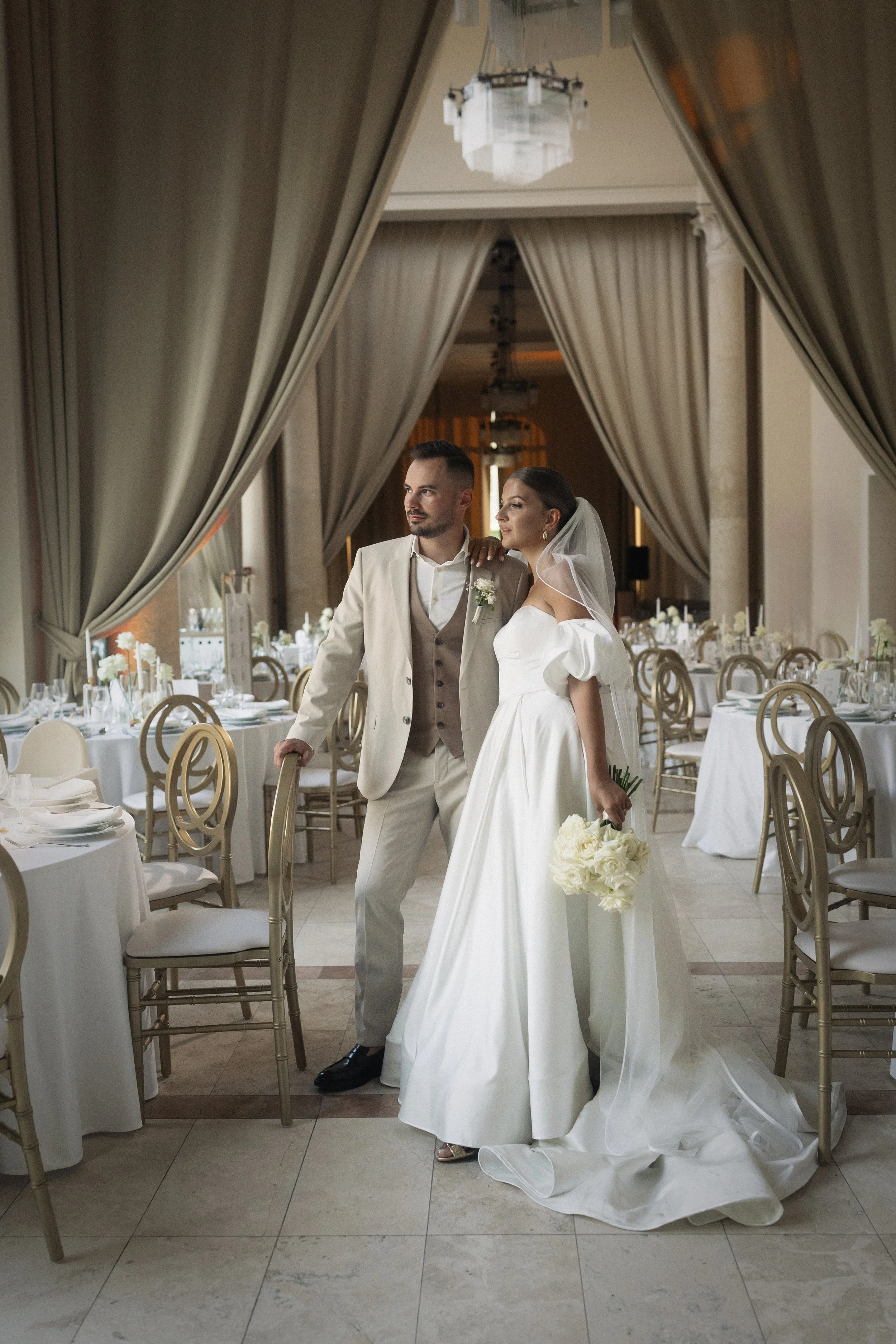 Modern fehér esküvői dekoráció a tatai Pálma Rendezvényházban - virágíves főasztal háttér, gyertyafényes asztaldekor és elegáns menyasszonyi pár