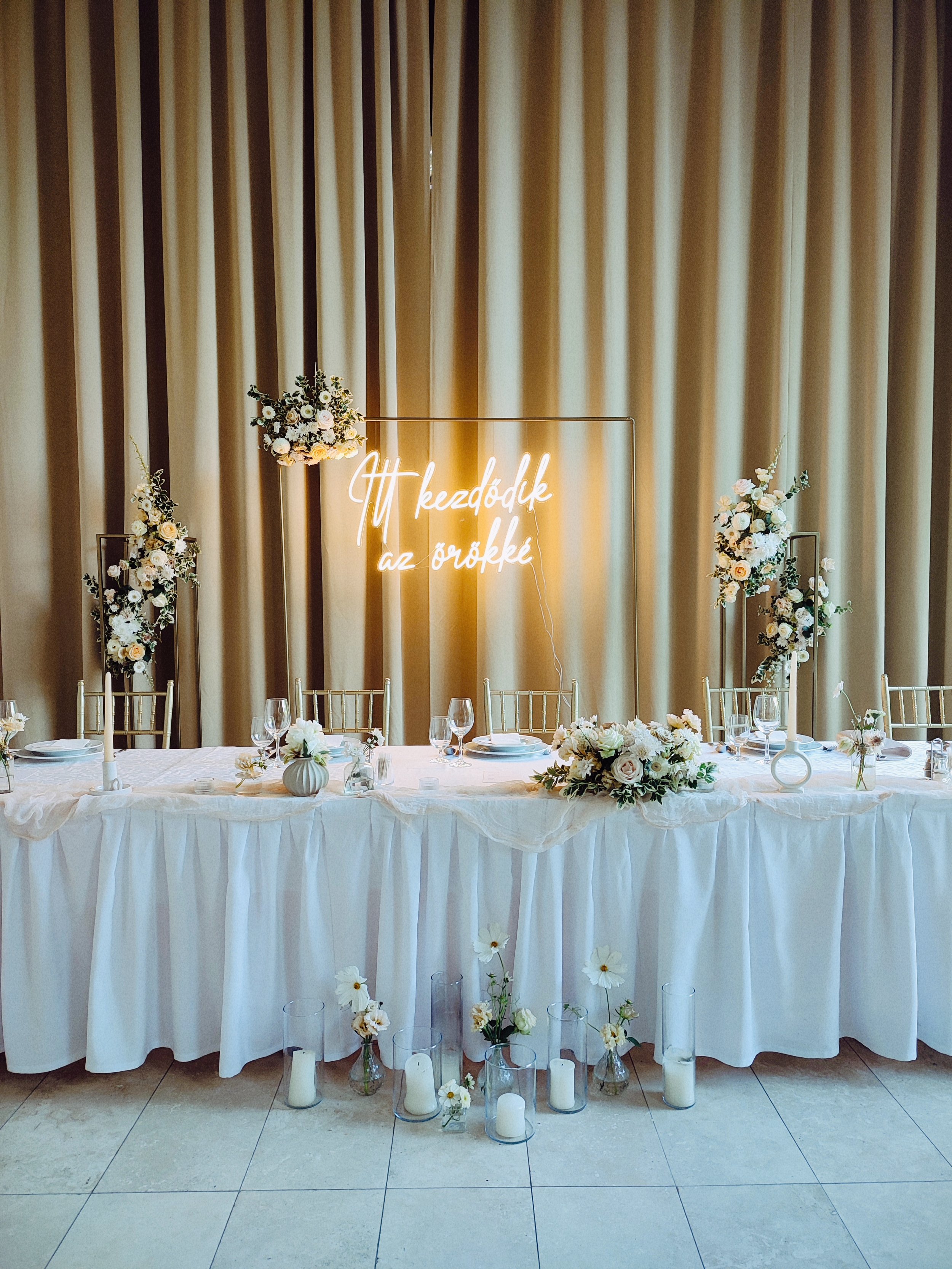 Elegáns esküvői főasztal fehér terítővel, virágdíszekkel, gyertyákkal és egy magyar neonfelirattal díszített háttérrel, arany székekkel és bézs függönyökkel a tatai Pálma Rendezvényházban.