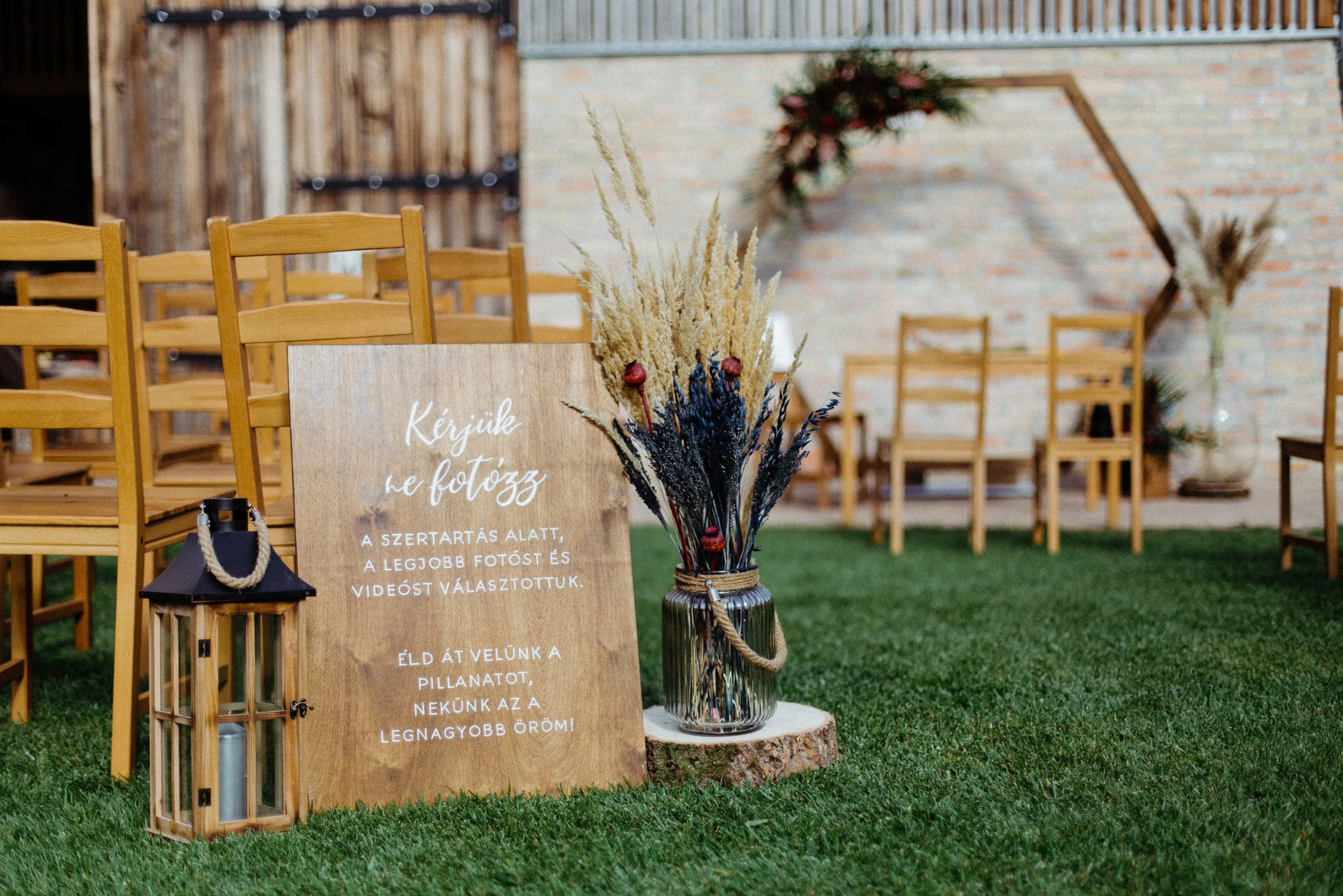 Egy fa tábla magyar felirattal, egy nagy üvegváza szárított virágokkal és egy lámpás a füvön egy feldíszített kültéri helyszínen, a háttérben székekkel és faldekorációkkal.