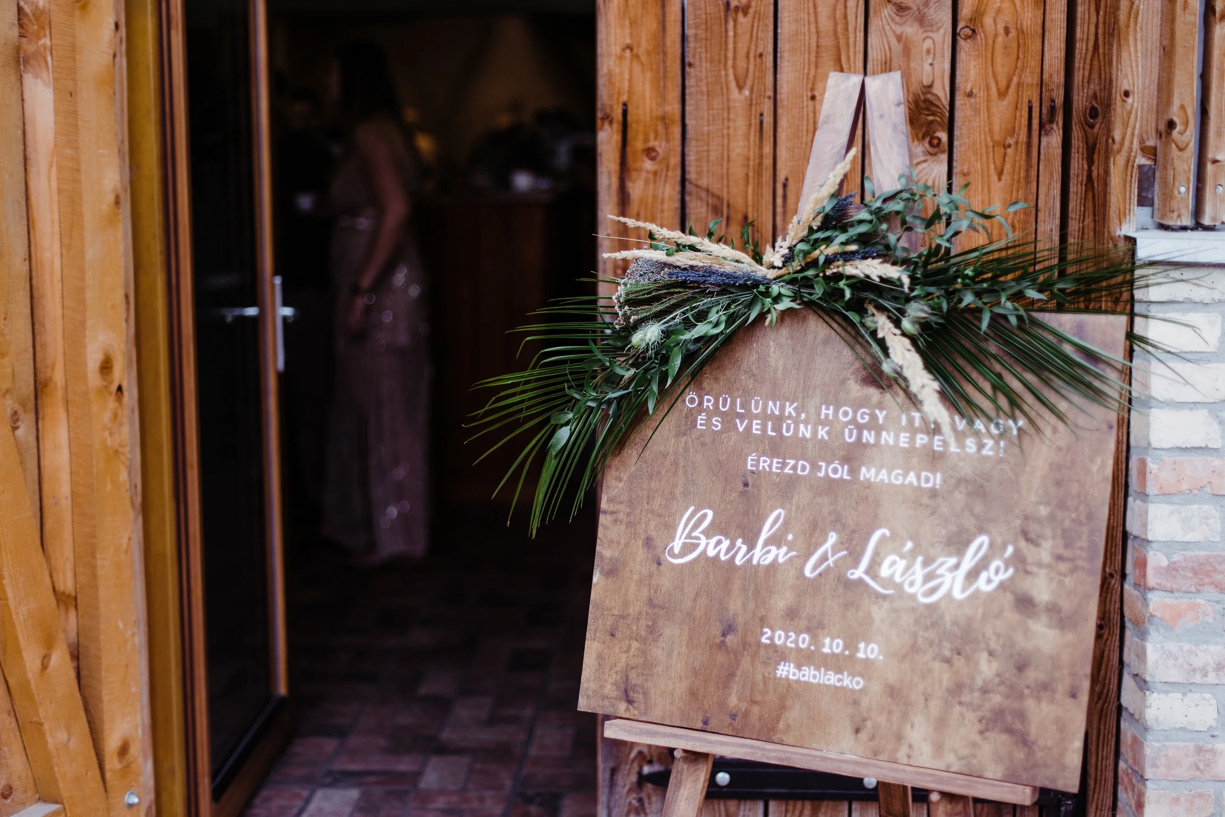 Esküvői üdvözlőtábla egy festőállványon, virágdíszítéssel egy fa állványon, magyarul köszönti a vendégeket.