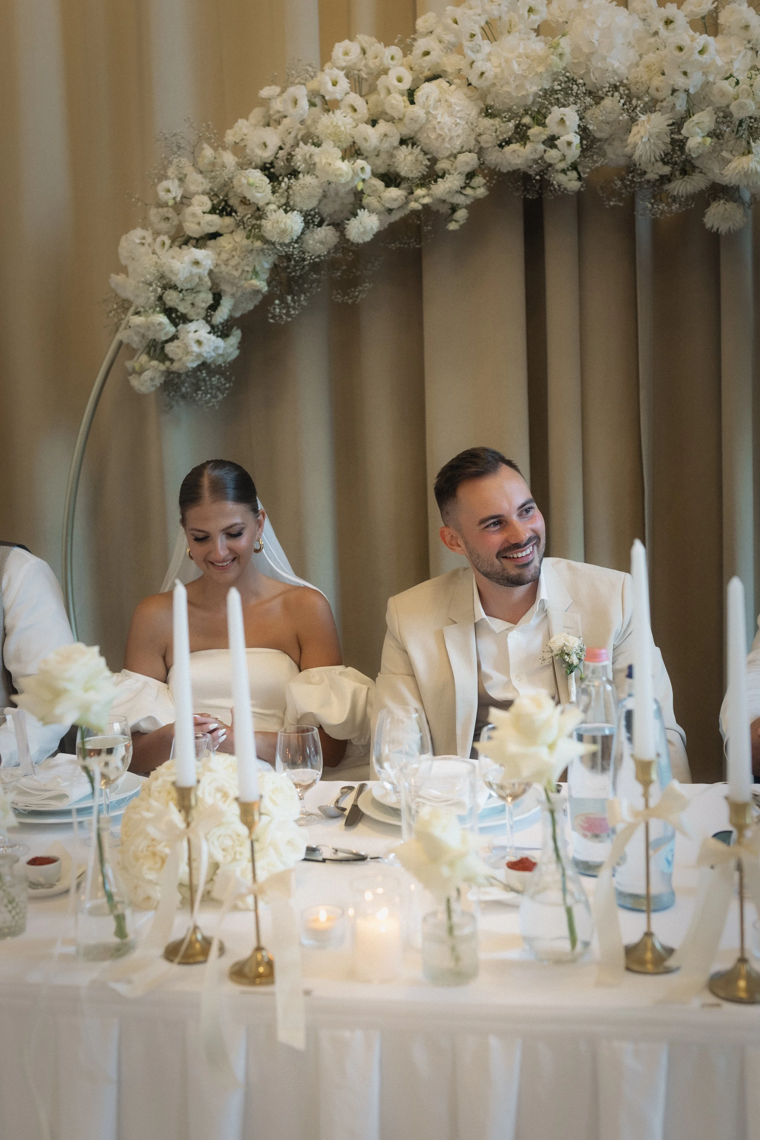 Menyasszony és vőlegény a főasztalnál fehér virágíves háttér előtt - elegáns esküvői dekoráció a Pálma Rendezvényházban