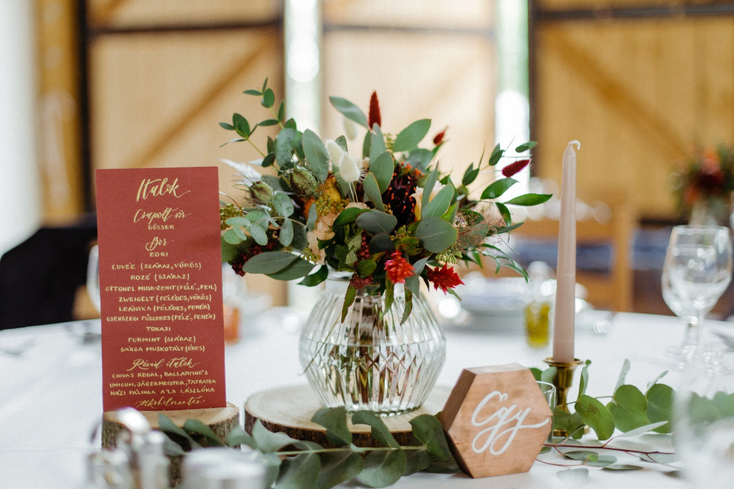 Esküvői vendégasztal dísze vegyes növényekkel és virágokkal díszített üvegvázával, magas rózsaszín gyertyával gyertyatartóban, piros étlappal és egy hatszögletű, fából készült asztalszámmal.
