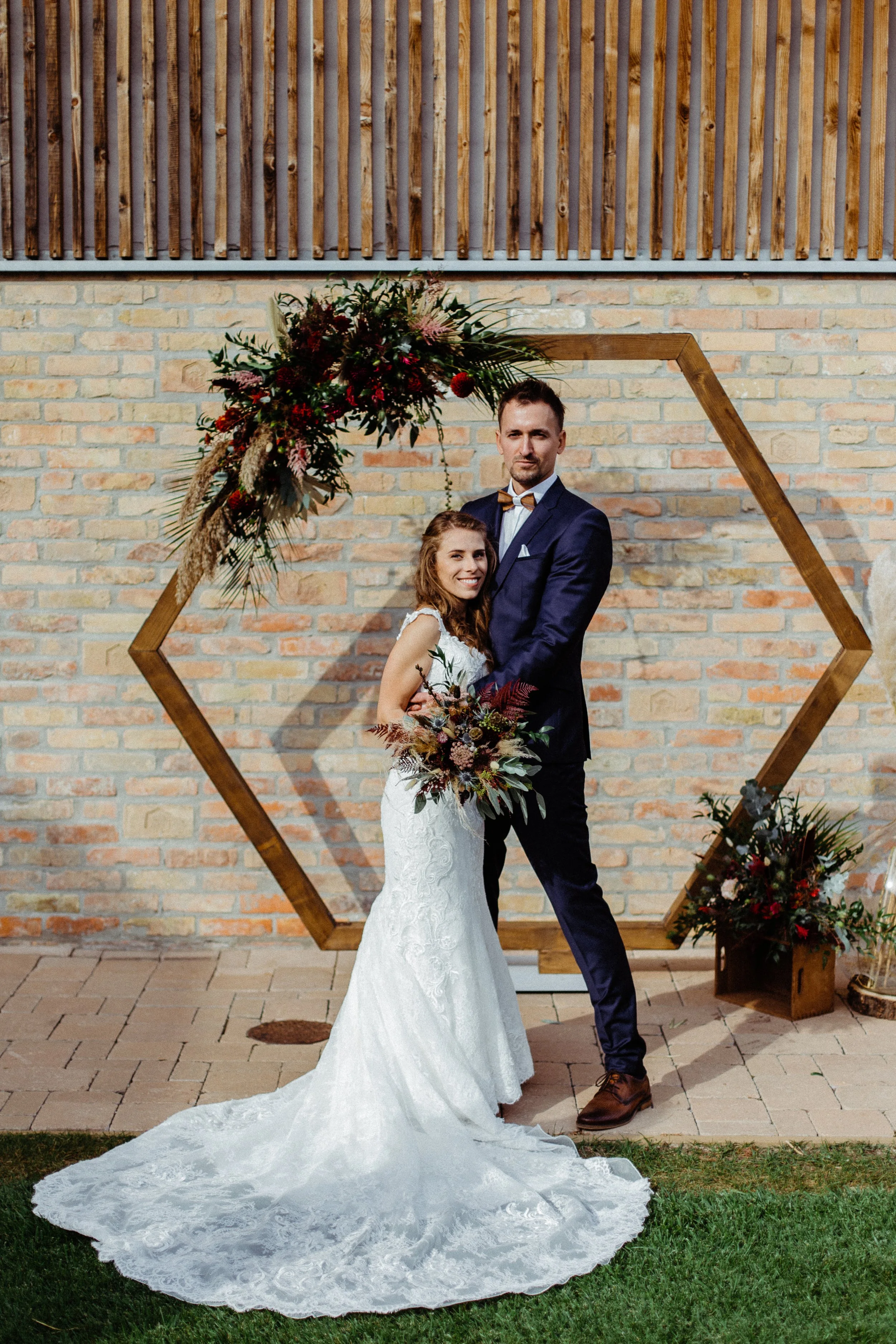 A menyasszony és a vőlegény együtt állnak egy díszes, hatszögletű, virágdíszekkel díszített, fából készült fakapu előtt.