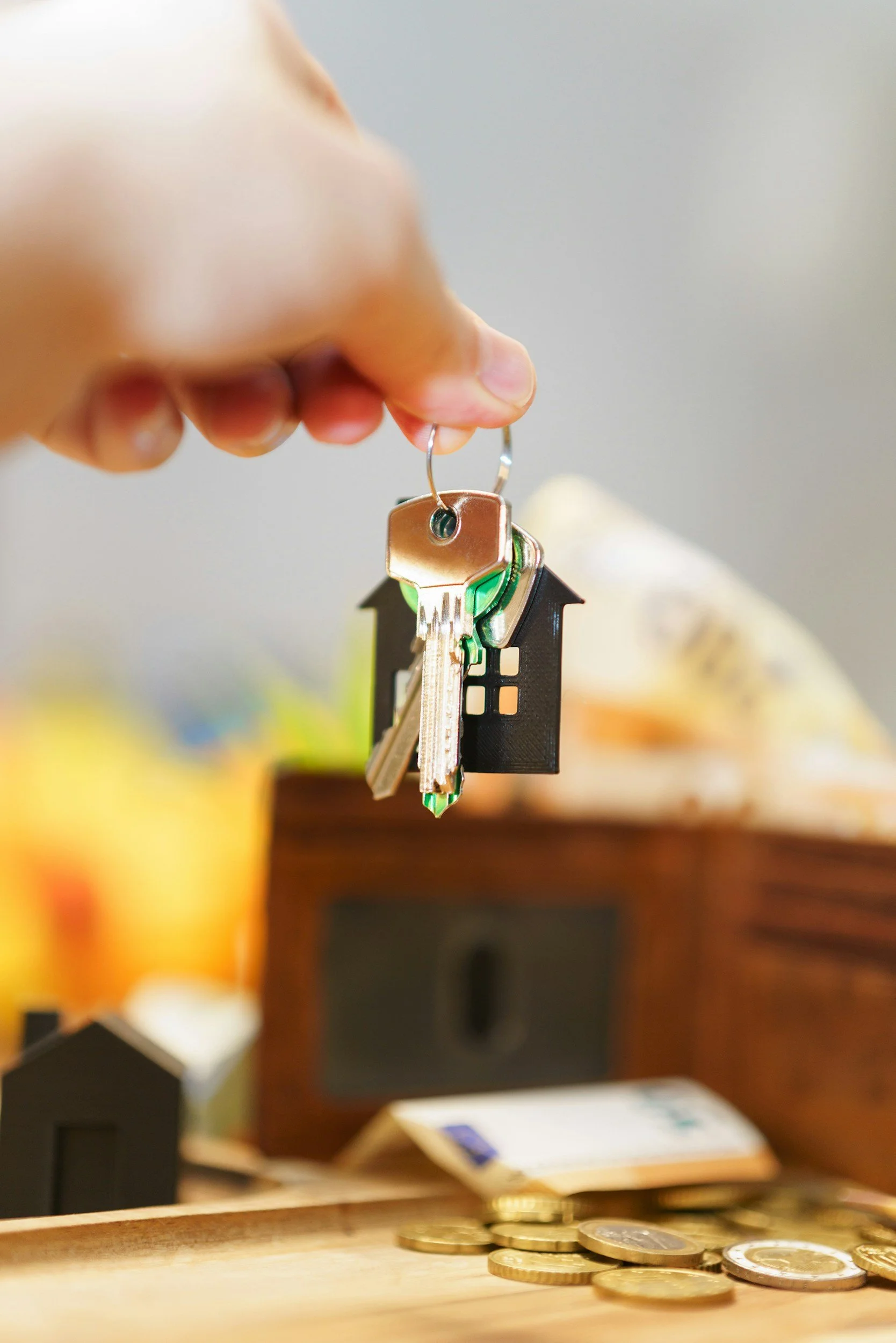 Close-up of a person's hand holding a set of keys with house-shaped keychains, hanging over scattered coins on a wooden surface, with a blurred background including furniture.