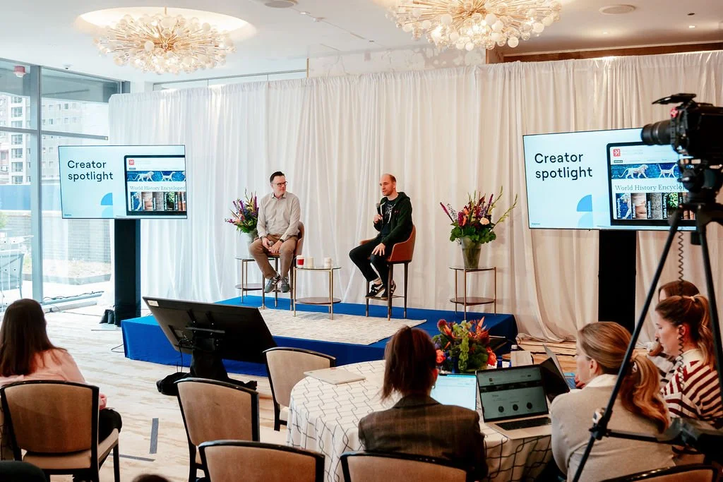 Two men on a stage during a speaker event at a conference room, with screens displaying 'Creator spotlight' and 'World History Encyclopedia', and an audience seated watching.