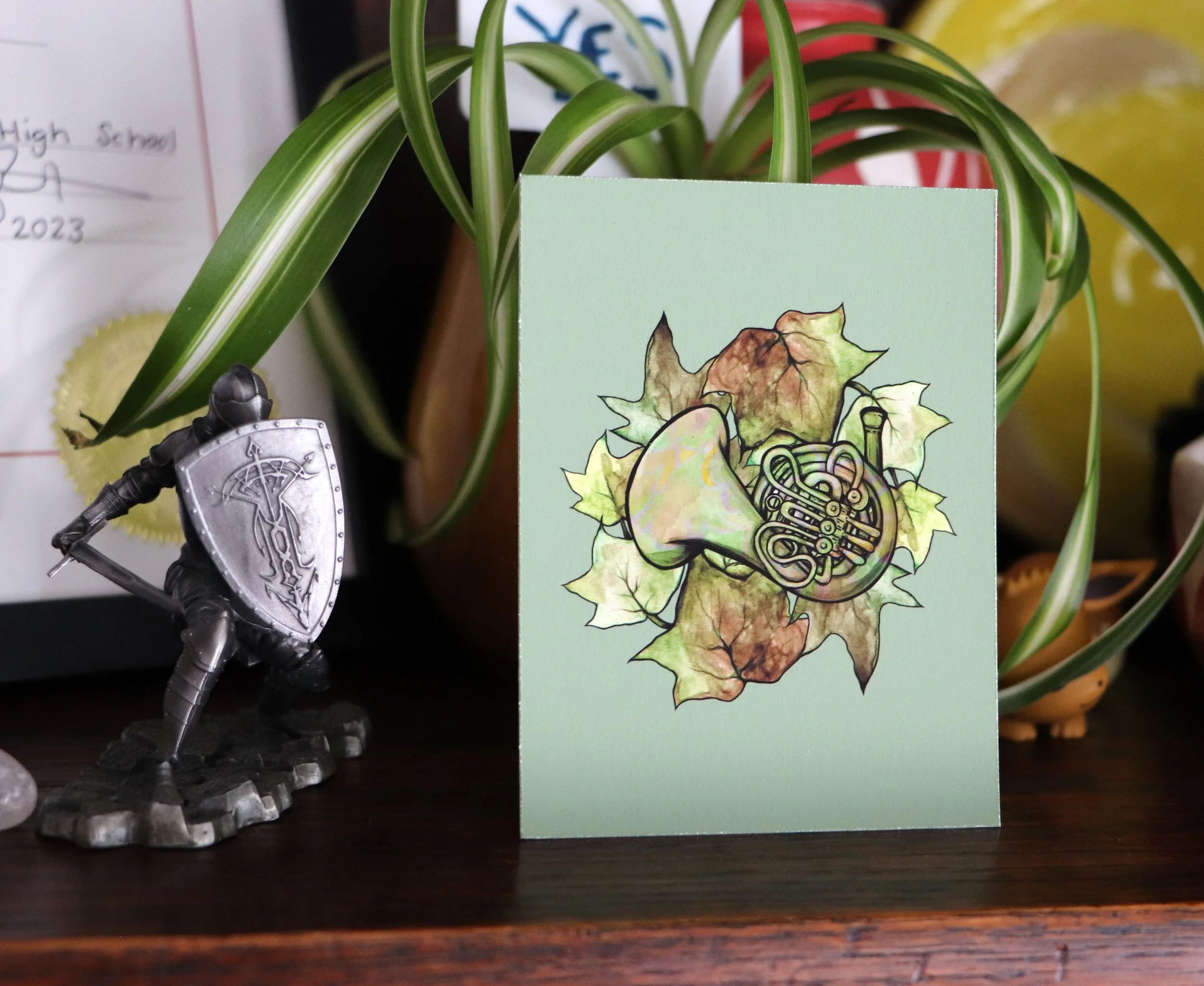A decorative card featuring an illustration of a French horn surrounded by autumn leaves, placed on a wooden surface with a potted plant and a knight figurine nearby.