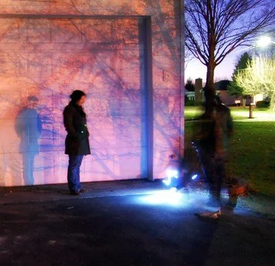Two people standing outside near a brick wall and a tree, with one person welding or working on something on the ground at night.