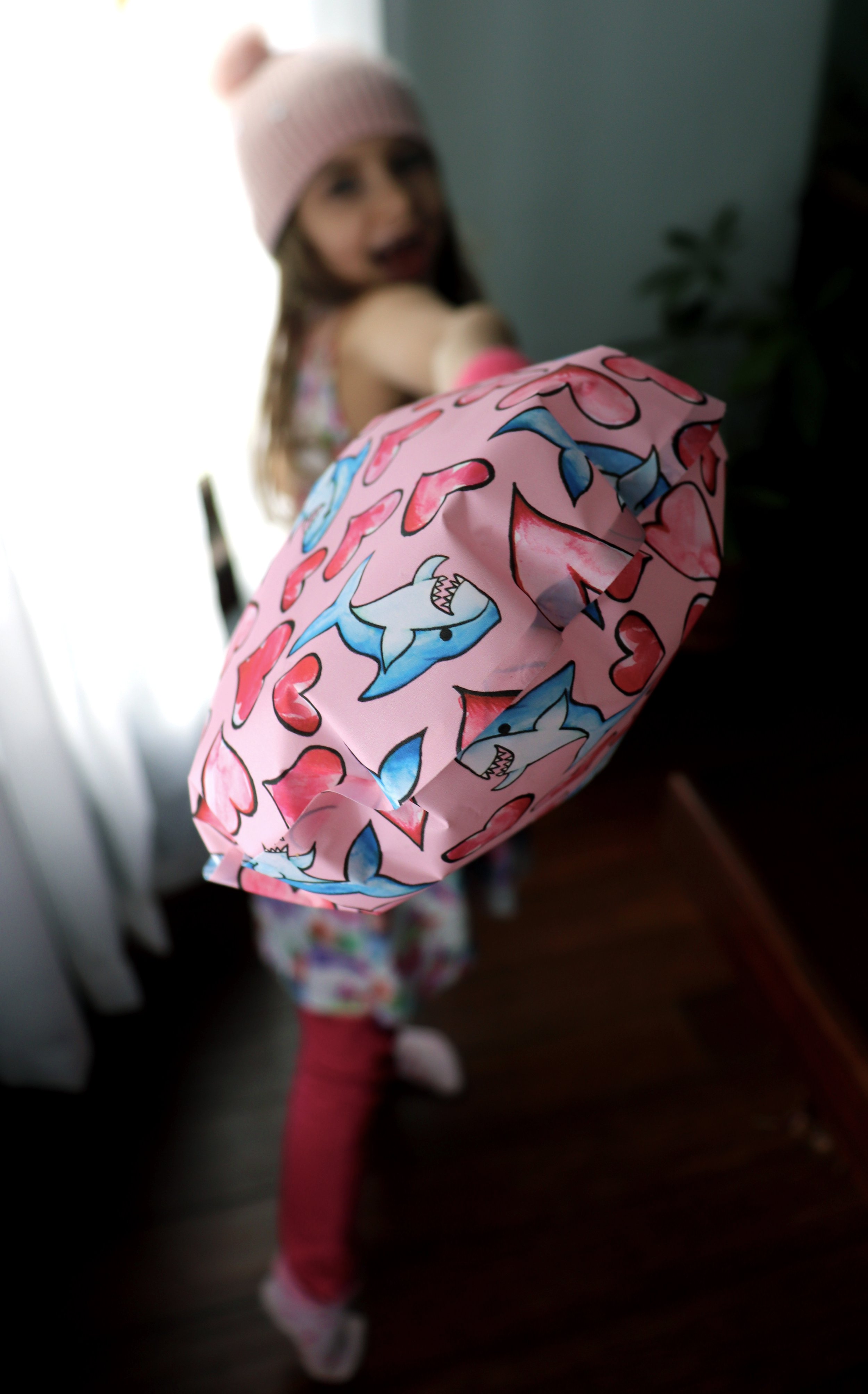 A young girl wearing a pink knit hat, patterned pajamas, and pink leggings is holding a pink gift bag with shark and heart designs. She is smiling and standing on a wooden floor inside a home.